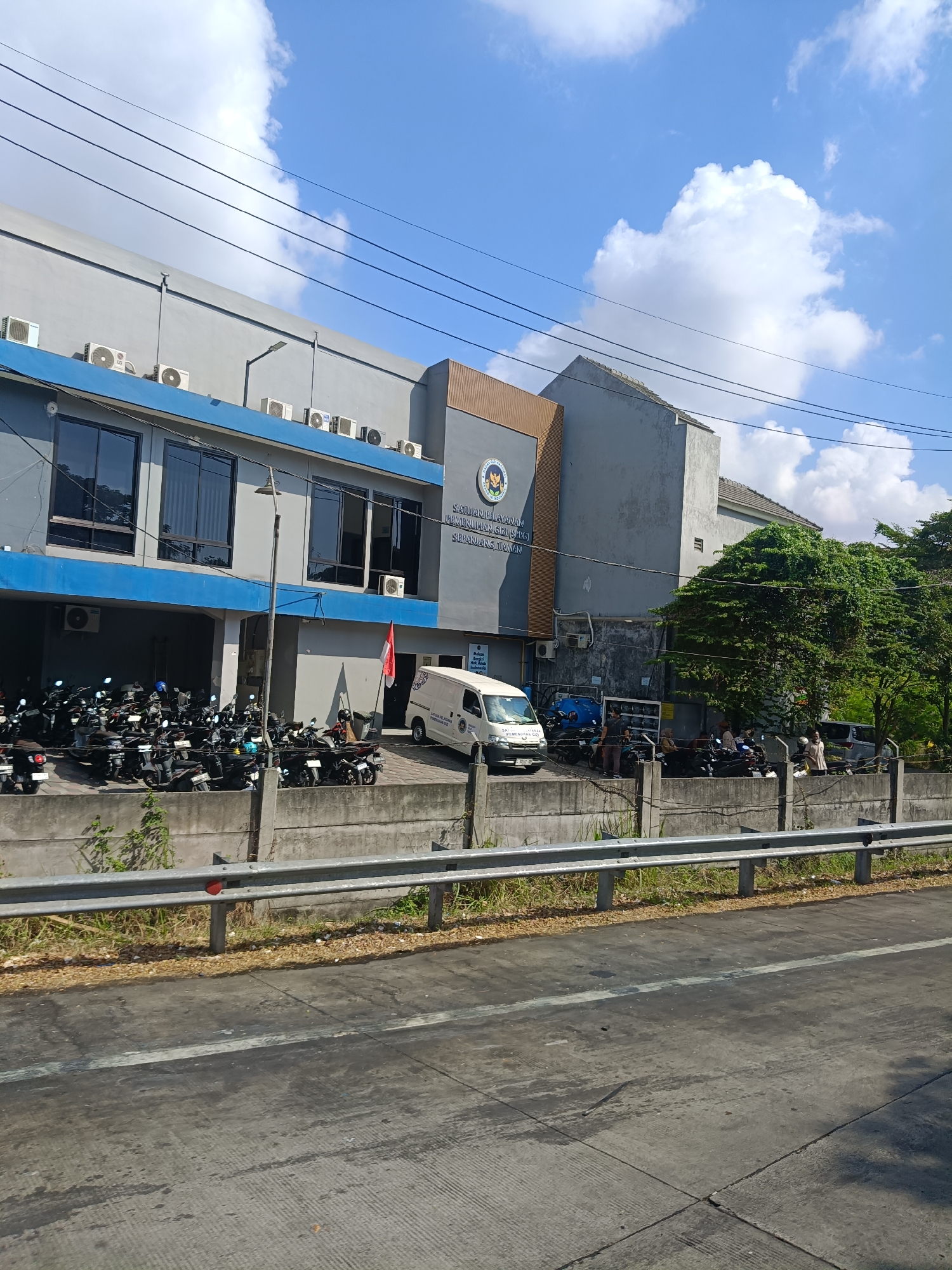 This image captures a street-level view of a building in Pagesangan, Indonesia, under a bright blue sky with scattered clouds. The building appears to be an official establishment, as indicated by the emblem on its facade and the text which reads "SATUAN PELAKSANA PEMERIKSAAN PENGUJIAN SERTIFIKASI & STANDARISASI". To the left of the building is a parking area filled with numerous motorcycles. A white van is parked in front of the building, and a small Indonesian flag is attached to its side.

In the foreground, a concrete barrier separates the street from the building's premises. The street itself is paved with asphalt, showing signs of wear and tear, and a white line marks a lane. The environment is characterized by urban infrastructure, with power lines visible against the sky. The presence of parked vehicles and a functioning building suggests a normal operational day.