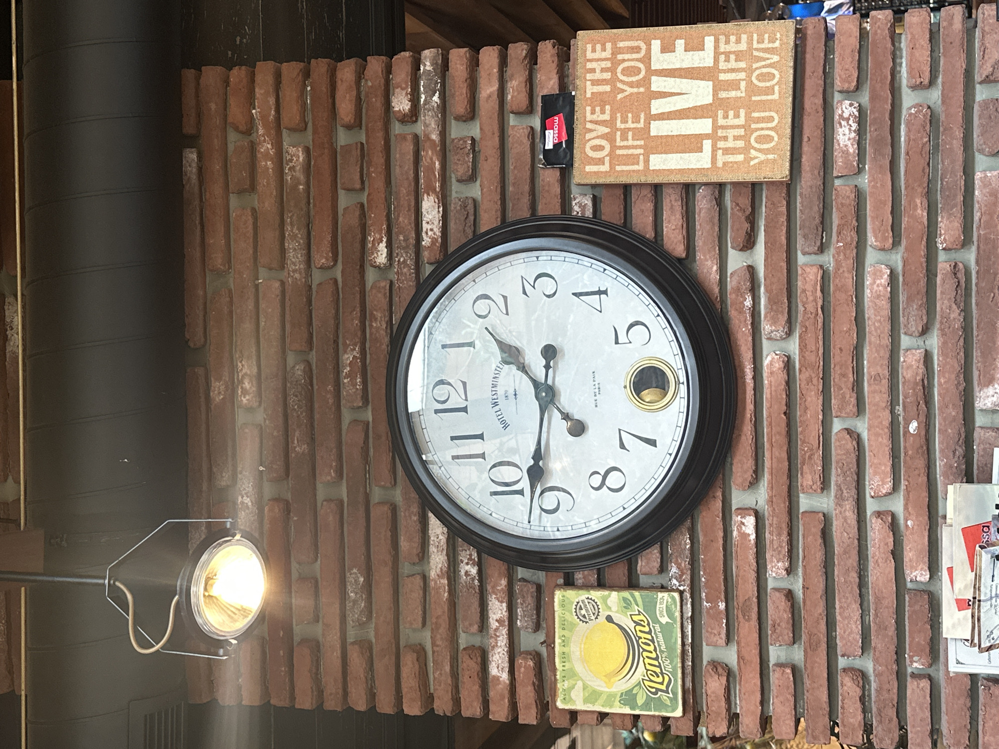 A close-up shot shows a section of a brick wall with several decorative items hanging on it. In the center is a large, round clock with a black frame. The clock face is white with black Roman numerals and ornate black hands. Below the clock face, a small pendulum swings. To the left of the clock is a small, square sign with a picture of a lemon and the text "Lemons 100% natural." To the right of the clock is a rectangular sign with an orange background and white text that reads "LOVE THE LIFE YOU LIVE THE LIFE YOU LOVE." A black and red packet is attached to the wall to the left of the orange sign. An industrial-style light fixture hangs from the ceiling above the clock. The brick wall appears aged and weathered, with some areas showing a white residue. The background above the brick wall consists of dark piping and wooden beams. The bottom of the image shows a stack of papers and some blurry objects. The context of the city of Varna, Bulgaria is not evident in the image.