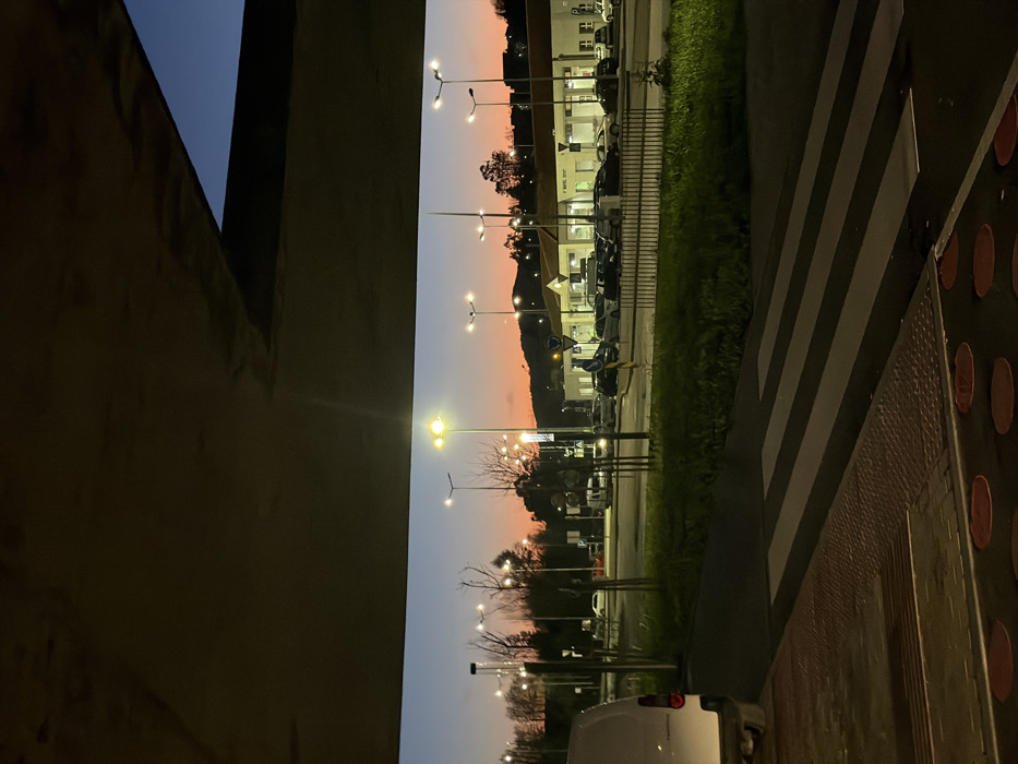 The image depicts an urban scene at dusk or early night in Penafiel, Portugal, characterized by artificial lighting against a twilight sky. The top half of the frame is dominated by the underside of a concrete structure, likely an overpass or a large architectural overhang.
In the immediate foreground, a white panel van is partially visible on the far left, showing its rear bumper and taillights. Adjacent to this, a pedestrian area features tactile paving with circular and linear raised patterns, leading to a clearly marked zebra crossing on a dark asphalt road.
The midground includes a multi-lane roadway bordered by a grassy median or verge. Numerous active streetlights, equipped with yellow-emitting lamps, illuminate the road and surrounding areas. Sparse, bare trees and shrubs are distributed across the landscape. A low metal fence separates the grassy area from a commercial parking lot.
In the background, a low-rise commercial building with a white facade and dark roofing is prominent. A sign on this building reads "P. NAFIEL," confirming the location in Penafiel. The parking lot in front of the building contains numerous parked vehicles of various types and colors. Several circular blue and white road signs are visible near the building's entrance. Behind the building, a prominent hill or elevated terrain, sparsely vegetated, is silhouetted against the sky, which transitions from a warm orange-pink at the horizon to a deeper blue overhead. No individuals are visible in the scene.