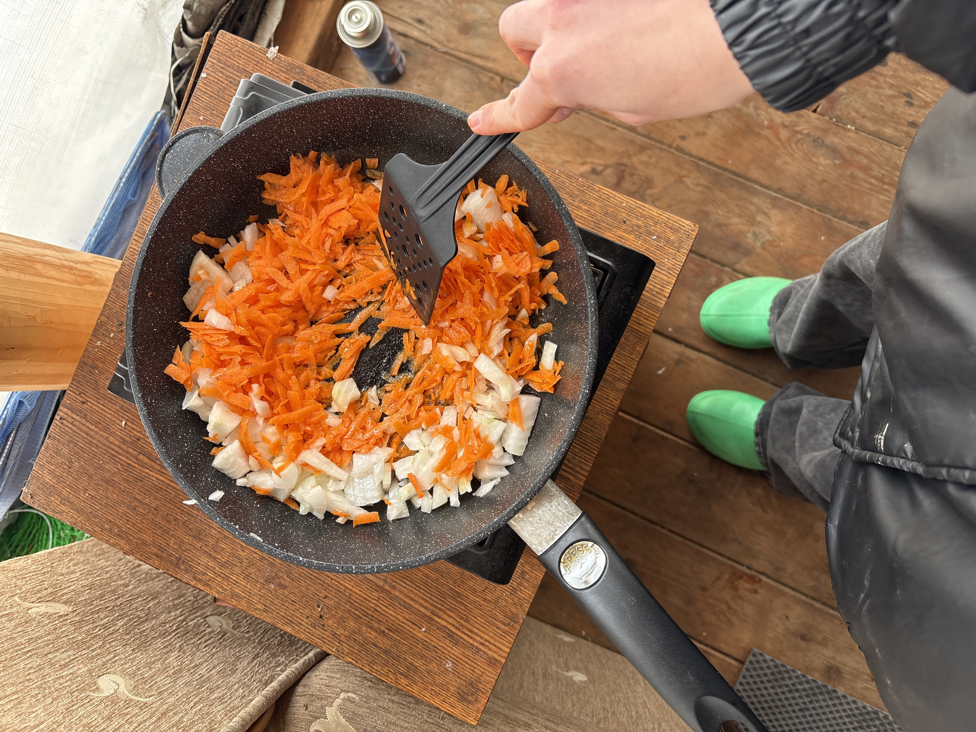 A high-angle shot depicts a person stirring a frying pan containing chopped carrots and onions on a wooden surface outdoors. The person is using a black slotted spatula. A small gas canister sits next to the pan. The individual is wearing a black jacket and grey trousers, with bright green rubber boots visible. The wooden surface appears to be a makeshift cooking station. The location is outdoors and potentially temporary. The exact city of Zheleznodorozhnyi, Russia cannot be determined from the image.