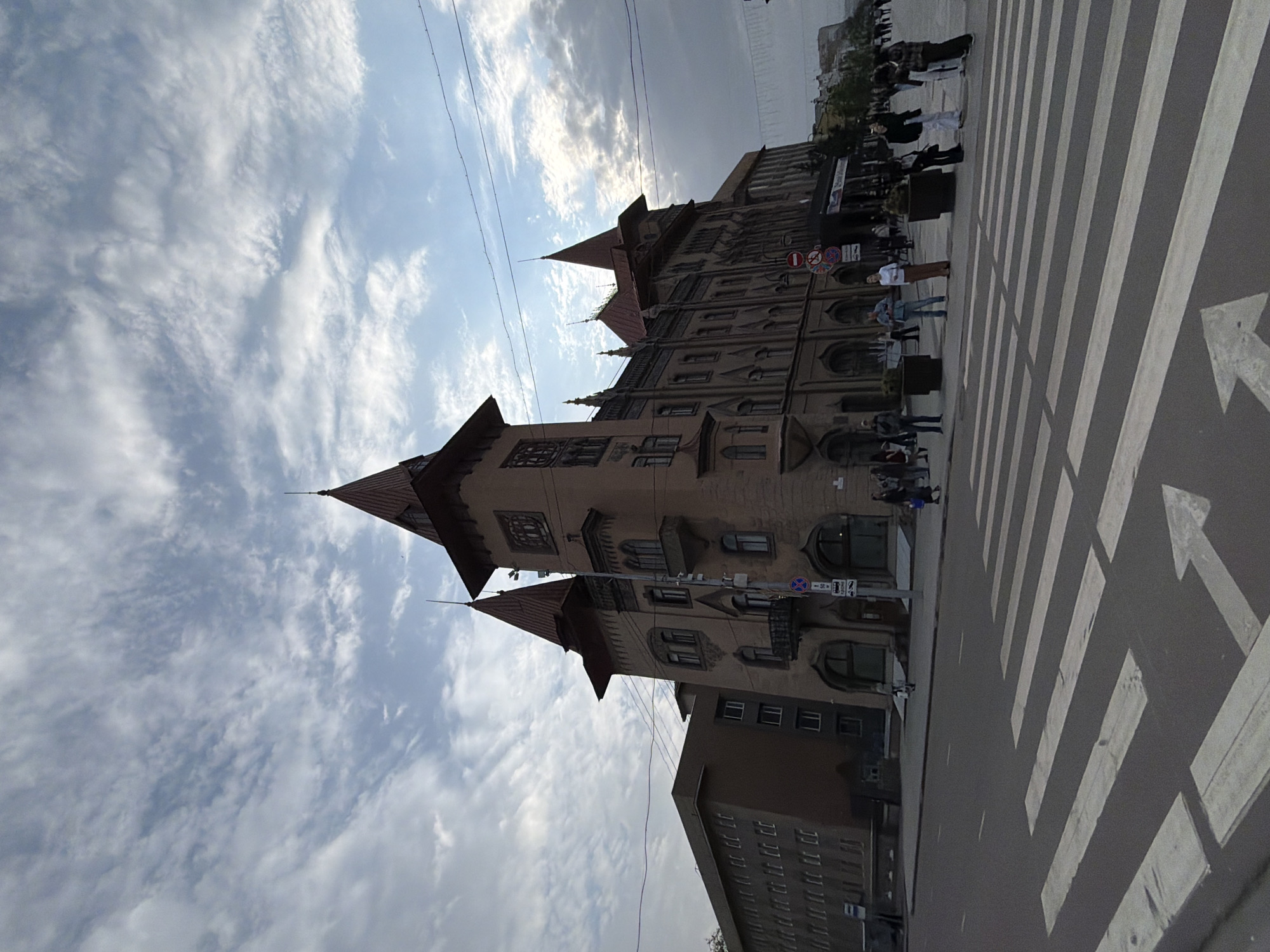 The image shows a building in Saratov, Russia. The building is multi-story and appears to be made of brick or stone, with a distinct architectural style featuring pointed roofs and arched windows. A crosswalk with white stripes and arrows is visible in the foreground. Several people are standing or walking near the building. The sky is partially cloudy. There are some overhead wires in the sky.