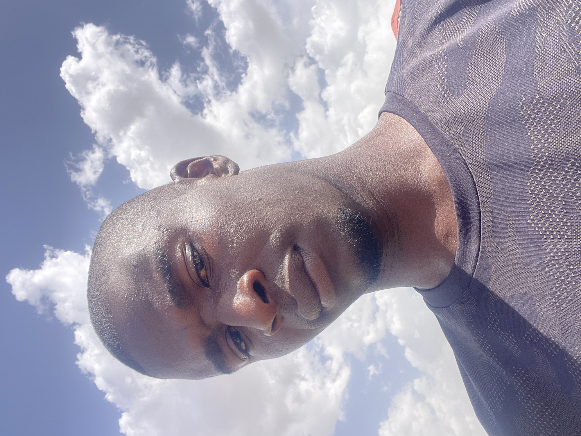 A close-up, angled selfie of a dark-skinned man with a shaved head is set against a backdrop of a blue sky with scattered white clouds. The man's face is slightly sweaty, and he is wearing a dark-colored t-shirt. No other people, subjects, objects, actions, interactions, or details about the environment suggest a specific location in Durimi, Nigeria.