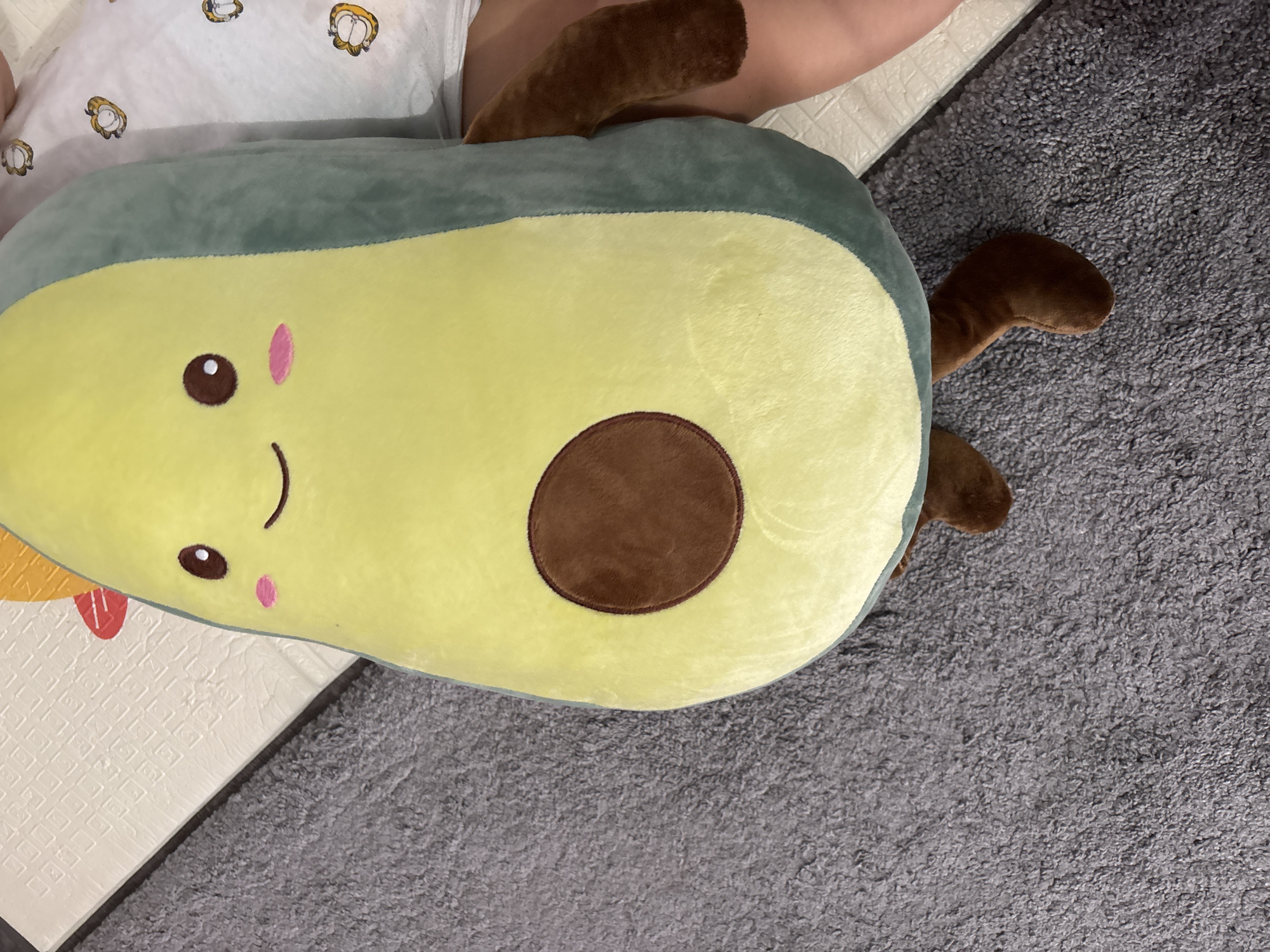 An overhead, medium shot captures a plush avocado toy lying on a textured grey carpet. The toy is large and appears to be filled with soft material. It is positioned diagonally, with its upper section on the left and lower section extending towards the right. The avocado has a friendly embroidered face with closed eyes, a simple curved mouth, and two pink circular blush marks. A prominent brown oval shape, representing the pit, is centered on the yellow part of the toy. Two short, brown limbs are visible, extending downwards from the lower part of the avocado. To the right of the toy, a portion of a person's legs and torso are visible, suggesting they are lying down next to it. The person is wearing light-colored clothing, possibly a t-shirt, with a faint pattern of cartoonish faces. The background also includes a section of a textured white mat, possibly a play mat, to the left of the carpet. The scene is indoor, with soft lighting. There is no indication of the city of Pernik, Bulgaria.
