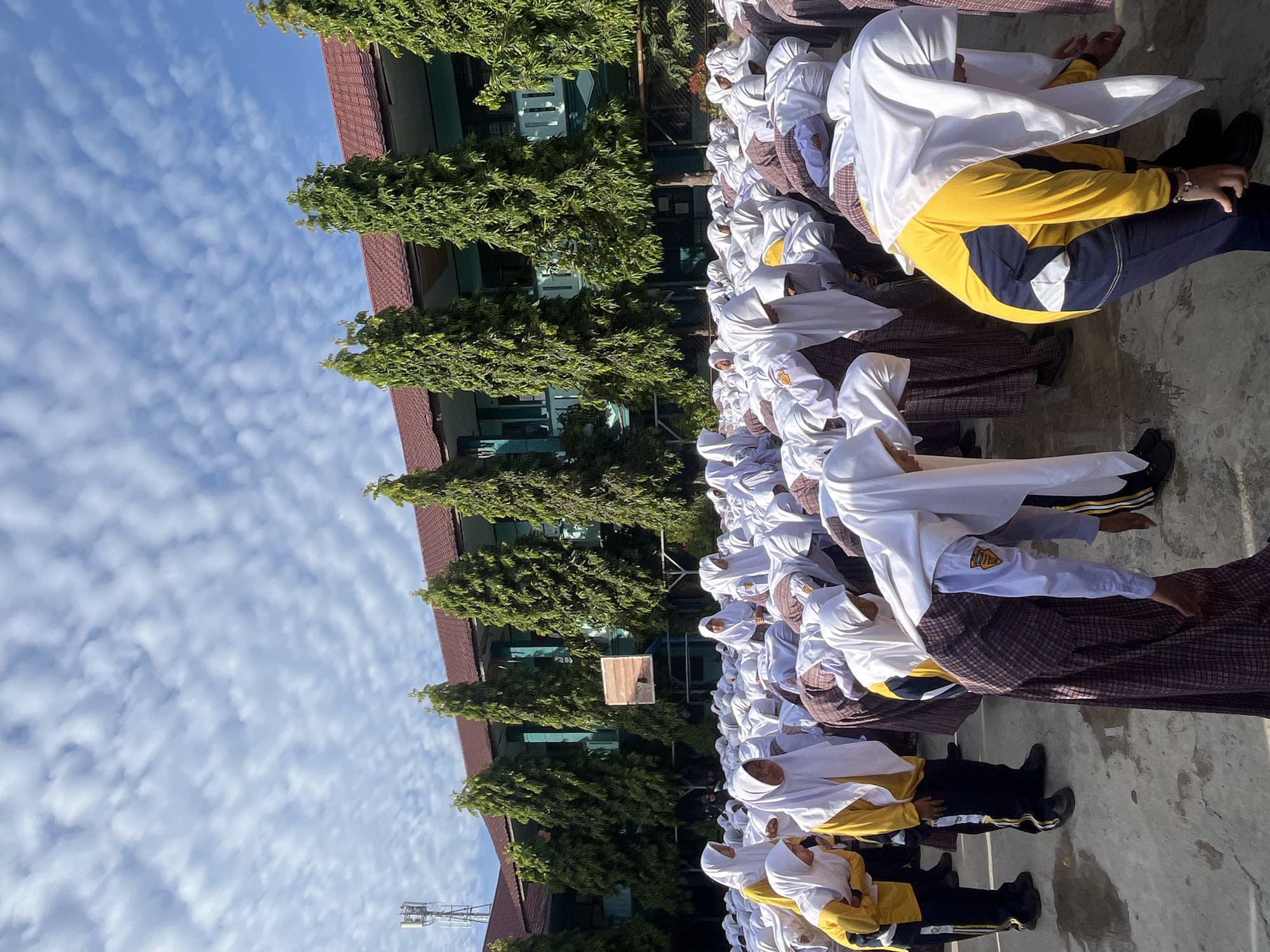 This image depicts a group of students, predominantly female, wearing white hijabs and school uniforms, engaged in what appears to be a physical exercise or a group activity outdoors. They are in a bent-over posture, facing downwards. The students are in a schoolyard, with a building in the background that has a red-tiled roof and green-painted walls. Several tall, slender trees are planted in front of the building, and a basketball hoop is visible in the middle ground. The ground is paved with concrete. The sky is blue with scattered white clouds, and a cell tower is visible in the distance to the left. This scene is likely taking place at a school in Lhokseumawe, Indonesia, given the architectural style and vegetation commonly found in the region.