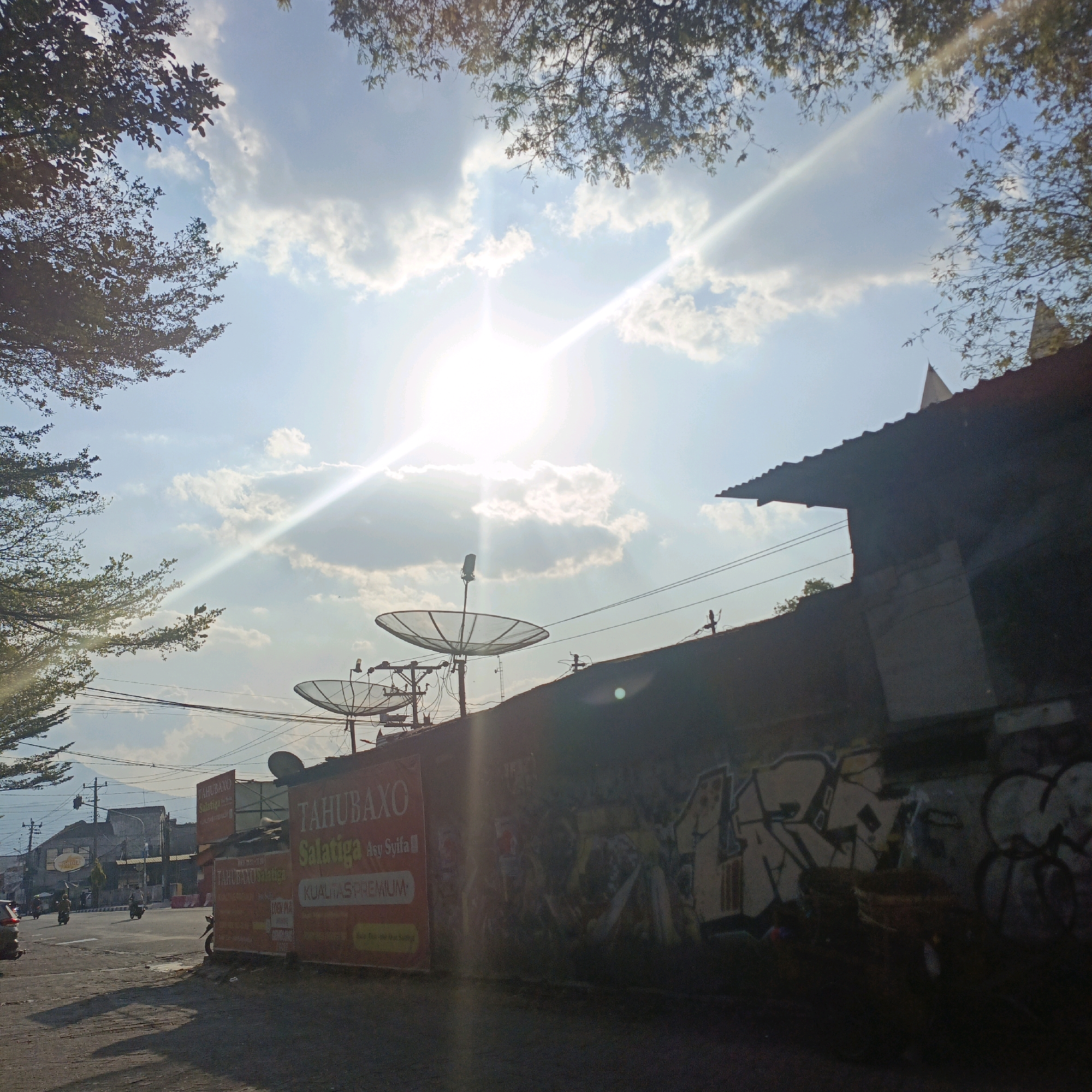 The image shows a street scene in Salatiga, Indonesia, bathed in bright sunlight with visible lens flare. The foreground features a dark, textured wall covered in graffiti, with a hint of a corrugated metal roofline above. To the left of the wall, a red and yellow advertisement is partially visible, featuring text that appears to be in Indonesian, possibly relating to food, as "TAHUBAXO" and "SALATIGA" are legible. Two satellite dishes are mounted on poles on top of the wall. Beyond the wall, a street is visible with traffic, including motorcycles and a car. The road curves towards buildings in the distance. Trees with sparse foliage frame the top of the image, with their branches silhouetted against a bright blue sky filled with fluffy white clouds. The overall impression is an urban setting on a sunny day.