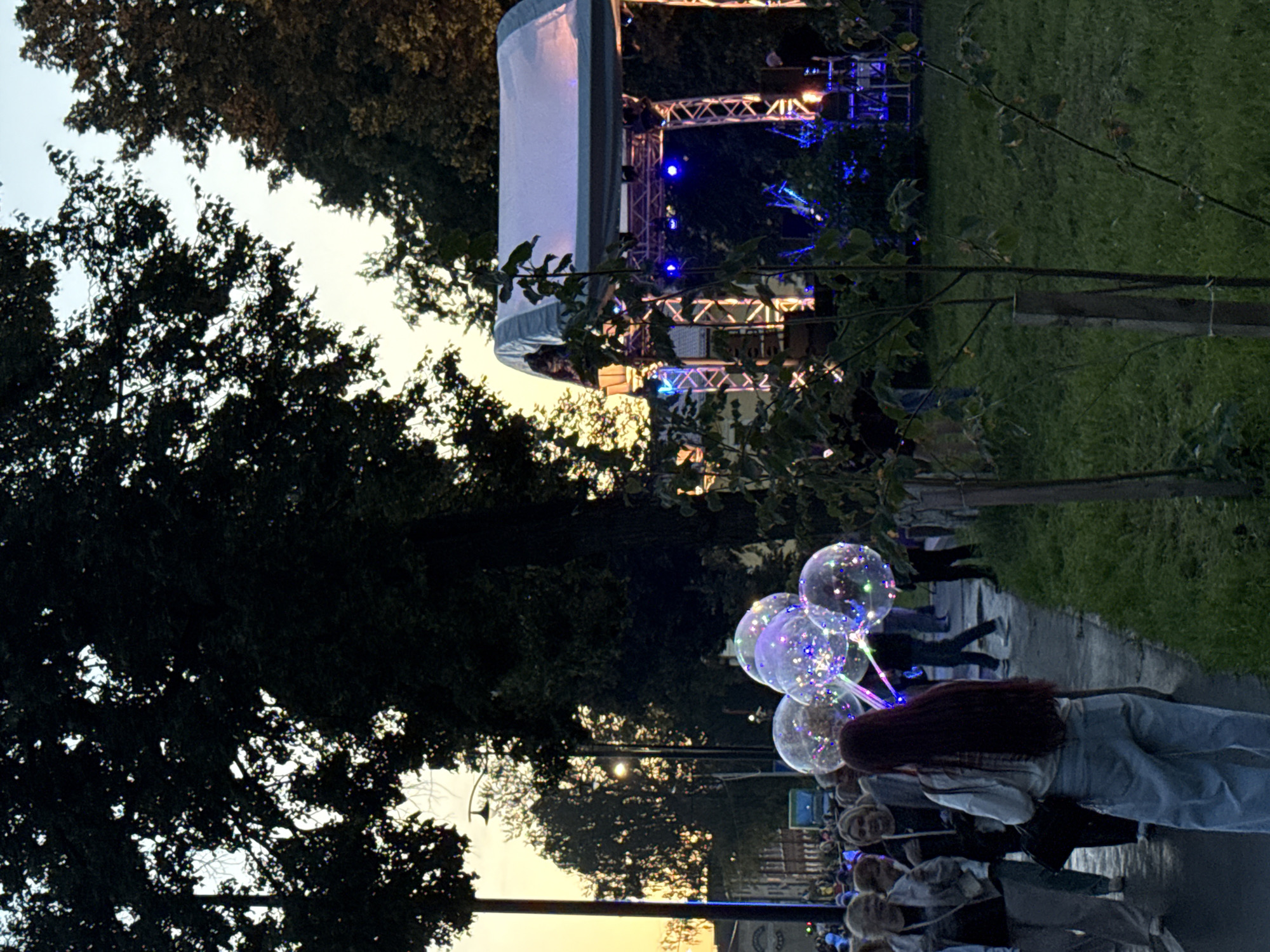 An outdoor stage with a white canopy is set up in a park in Votkinsk, Russia, at dusk. The stage is illuminated with blue and yellow lights, and there are speakers and other equipment visible. A crowd of people is gathered in front of the stage, some holding illuminated balloons. The foreground shows a pathway with grass on either side. The trees in the background are dark silhouettes against the sky.