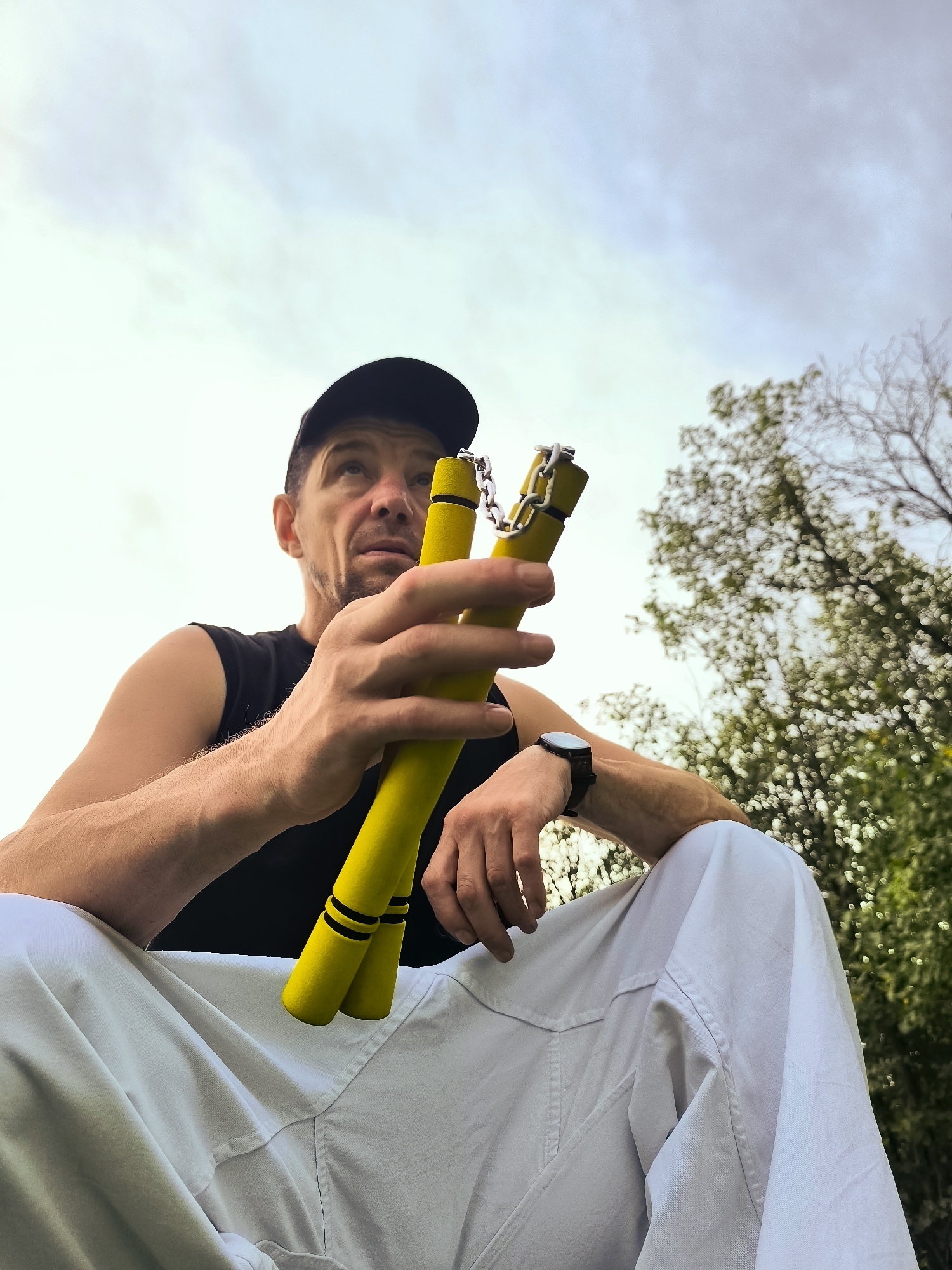 A low-angle, medium shot shows a man wearing a black baseball cap and a black sleeveless shirt, holding yellow nunchaku with a metal chain in his right hand. He is seated outdoors, with his legs crossed and covered in white pants. The background consists of a cloudy sky and blurred trees, suggesting a natural or park setting, not within the city administration of Qarağandı, Kazakhstan. The man's left arm is resting on his knee. A black wristwatch is visible on his left wrist.