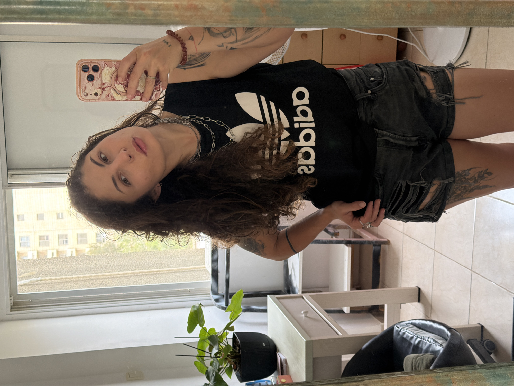 The image shows a person with long, curly brown hair, wearing a black Adidas t-shirt and ripped black denim shorts, taking a selfie in a mirror. The phone case is pink with a floral design. The person is standing indoors, possibly in an apartment. Visible in the background are a window with a view of a building across the street, a plant in a black pot, and other furniture. The window suggests a location in an urban environment. Based on the information provided, this location is identified as Kiryat Bialik, Israel.