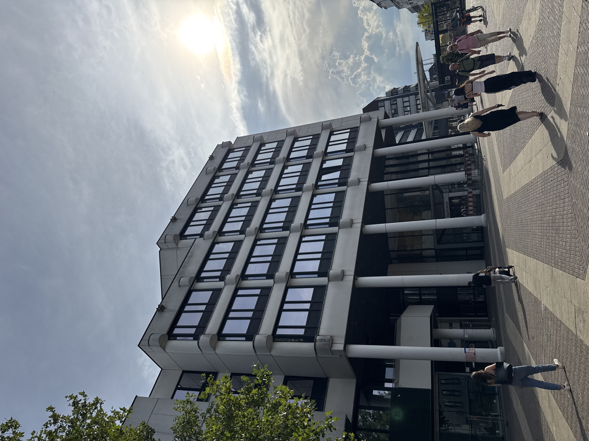 The image depicts the Rathaus (City Hall) in Rheine, Germany, on a partly cloudy day with the sun breaking through. The modern, multi-story building features a concrete facade with numerous dark-framed windows and is supported by large, white cylindrical columns at its base. A pedestrian walkway with a brick pattern extends in front of the building, leading towards the entrance.
Several individuals are visible in the foreground and midground. In the foreground, a woman in jeans and a black top walks away from the camera. Further back, a group of four people walks across the plaza, with two women in dresses in the lead, followed by two men. To the right, another group of people is present, one of whom is on a bicycle. In the midground, a man is seen pushing a stroller. Construction barriers are visible near the entrance of the Rathaus. Trees with green foliage are present on the left side of the image. The sky is filled with a mix of bright sun and scattered clouds.