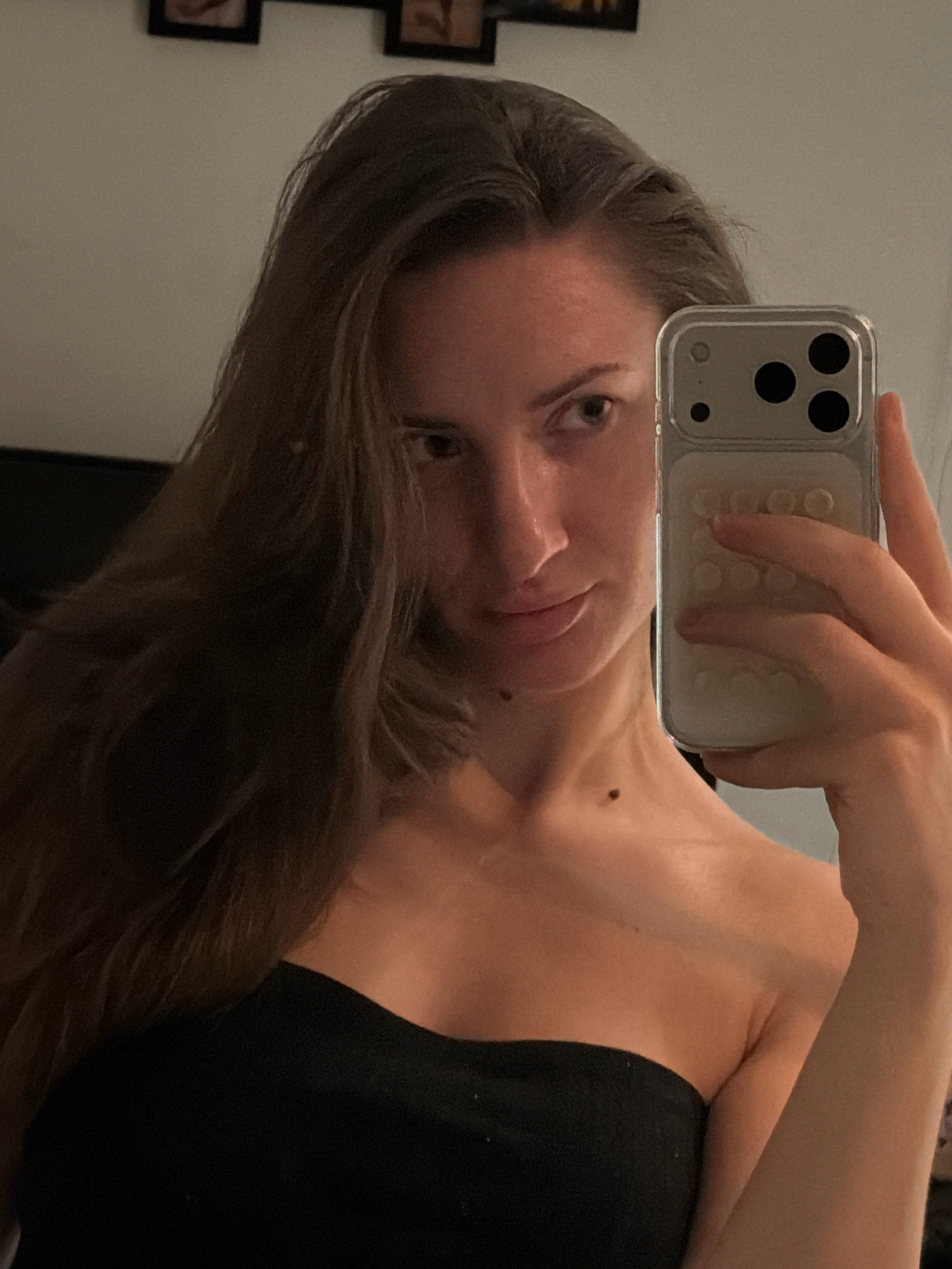 A woman takes a mirror selfie in what appears to be a dimly lit room. She is wearing a black strapless top. She has long, brown hair that is partially obscuring her face. She is holding a smartphone in her right hand, with the camera facing the mirror. The phone case has a textured pattern of circular indentations. The background of the image is a plain wall. There are framed pictures on the wall in the upper left portion of the image, suggesting a domestic setting, potentially in Sharm El Sheikh, Egypt.