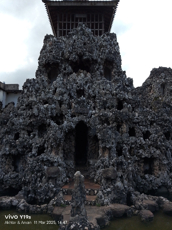 The image shows a large, rock-like structure with a wooden roof, located at the coordinates -6.736, 108.544, which correspond to the Keraton Kasepuhan Palace in Cirebon, Indonesia. The structure appears to be constructed from a rough, gray material, possibly concrete or stone, with numerous openings and alcoves of varying sizes. The roof is a simple structure with a gabled shape and latticework panels. It is unclear what the purpose of the structure is, but it is likely part of a larger complex, possibly a shrine or a monument. The surrounding area is relatively empty, with a small body of water in the foreground and a portion of another rocky structure in the background. The scene is captured from a low angle, giving the impression of being quite tall. A small sign can be seen on the wall above a doorway with "KEPITEN" written on it. The image appears to be taken in daylight, but the overcast sky suggests it may have been cloudy. The overall atmosphere is peaceful and serene.