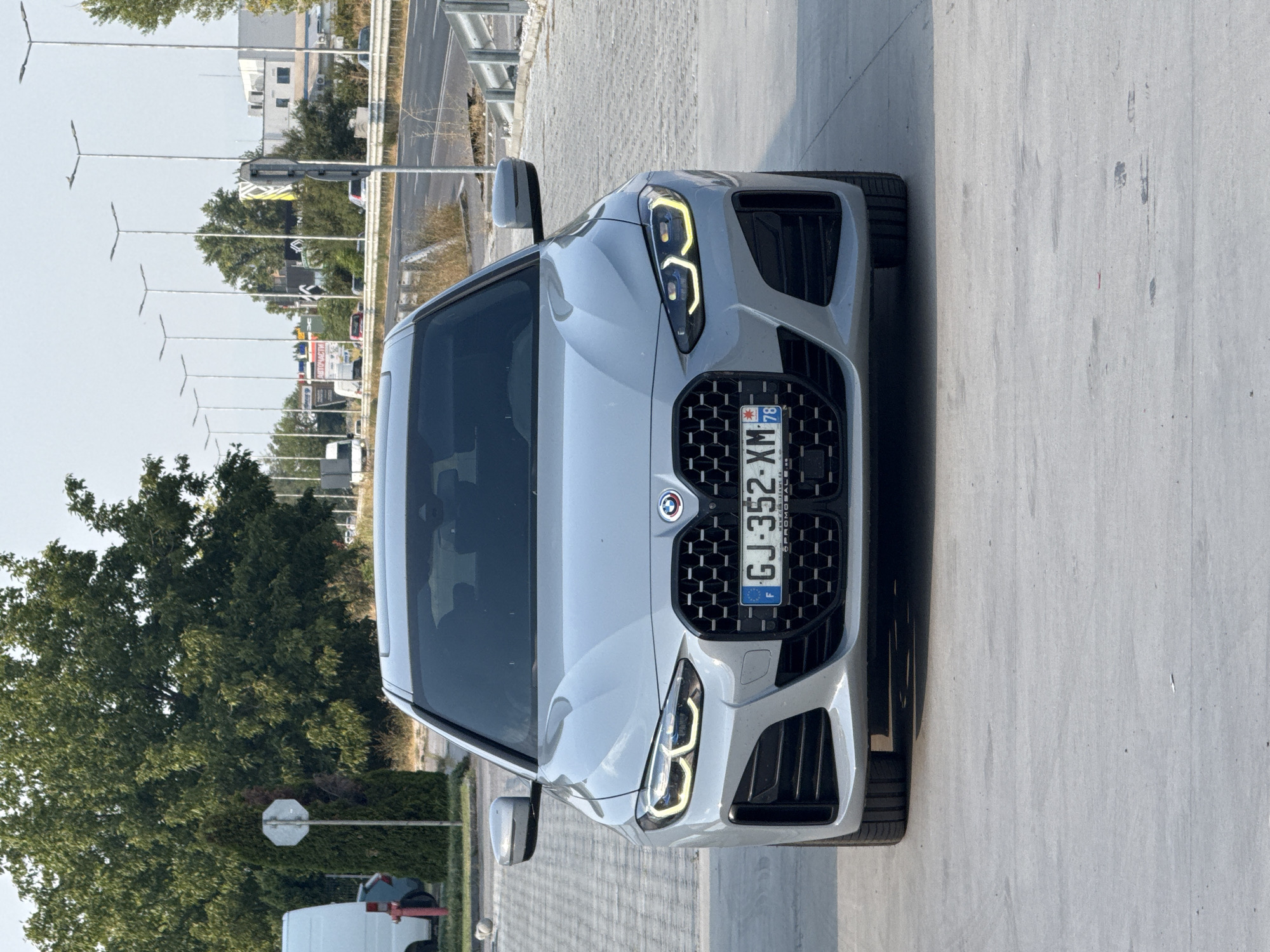 The image shows the front of a silver BMW sedan parked on a paved lot. The car's license plate reads "GJ 352 XM" with a French flag emblem and the number 78. In the background, there is a road with passing vehicles, streetlights, and buildings. Trees and a white van are also visible. The scene appears to be an outdoor commercial or industrial area, possibly near a roadside. The location is identified as Plovdiv, Bulgaria.