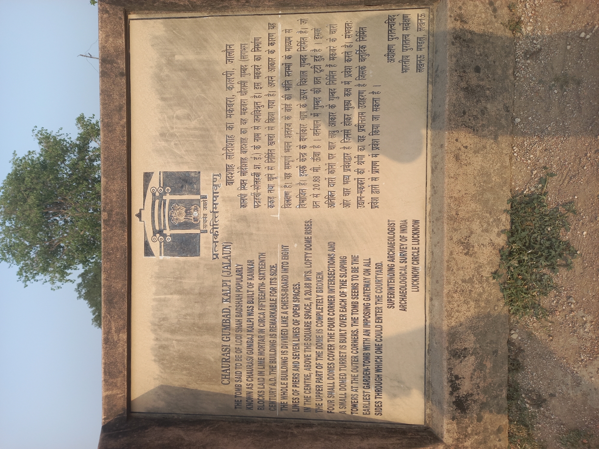 The image depicts a stone informational sign with text in both English and Hindi. The sign describes the Chaurasi Gumbad, located in Kalpi (Jalaun), India. The English portion of the text states: The tomb is said to be of Lodi Shah Badshah popularly known as Chaurasi Gumbaj Kalpi was built of Kankar blocks laid in lime mortar in circa fifteenth-sixteenth century A.D. The building is remarkable for its size. The whole building is divided like a chess board into eight lines of piers and seven lines of open spaces. In the centre, above the square space, a 20.88 mts. lofty dome rises. The upper part of the dome is completely broken. Four small domes cover the four corner intersections and a small domed turret is built over each of the sloping towers at the outer corners. The tomb seems to be the earliest garden-tomb with an imposing gateway on all sides through which one could enter the courtyard. The sign also indicates that the Superintending Archeologist of the Archeological Survey of India, Lucknow Circle Lucknow, put the sign together.
Above the text is the emblem of India, and the entire sign is set in a stone frame. A tree is visible in the upper left corner, suggesting an outdoor environment, and gravel is on the ground to the right side of the sign.
