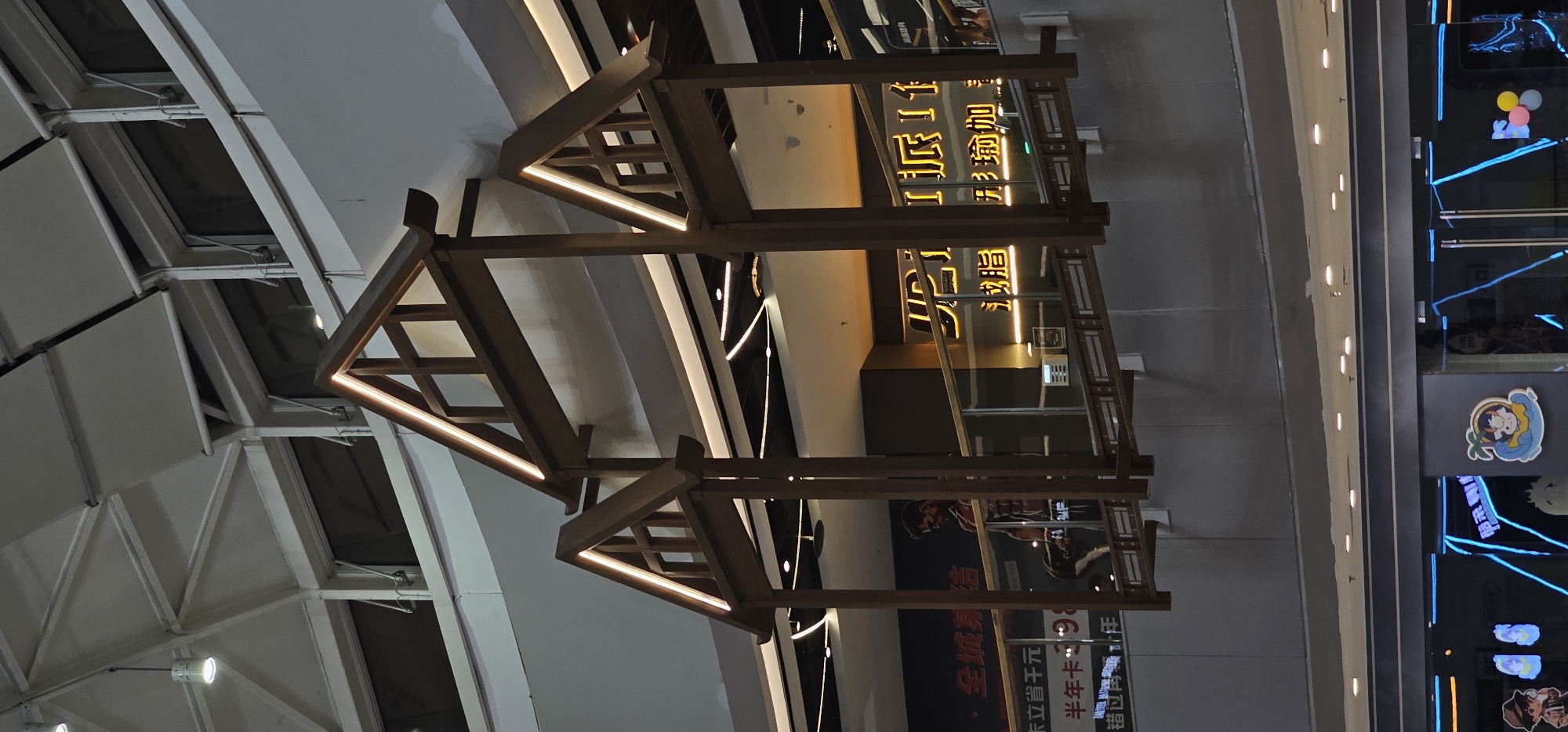 The image shows the interior of a commercial building, likely a shopping mall or an entertainment complex, in Shapingba District, China. In the foreground, a wooden architectural structure with a pitched roof design, illuminated by integrated LED strip lights, is suspended in the air. Below this structure, a glass partition separates two levels of the building. Through the partition, a large advertisement featuring a muscular man holding a barbell is visible, along with text in Chinese characters. To the right of this advertisement, another sign with illuminated Chinese characters can be seen. The background reveals the building's ceiling with a white, angular structure and multiple light fixtures. The lower part of the image shows a storefront with blue neon lighting and cartoon-like decals. The overall scene depicts a modern commercial environment with decorative architectural elements.