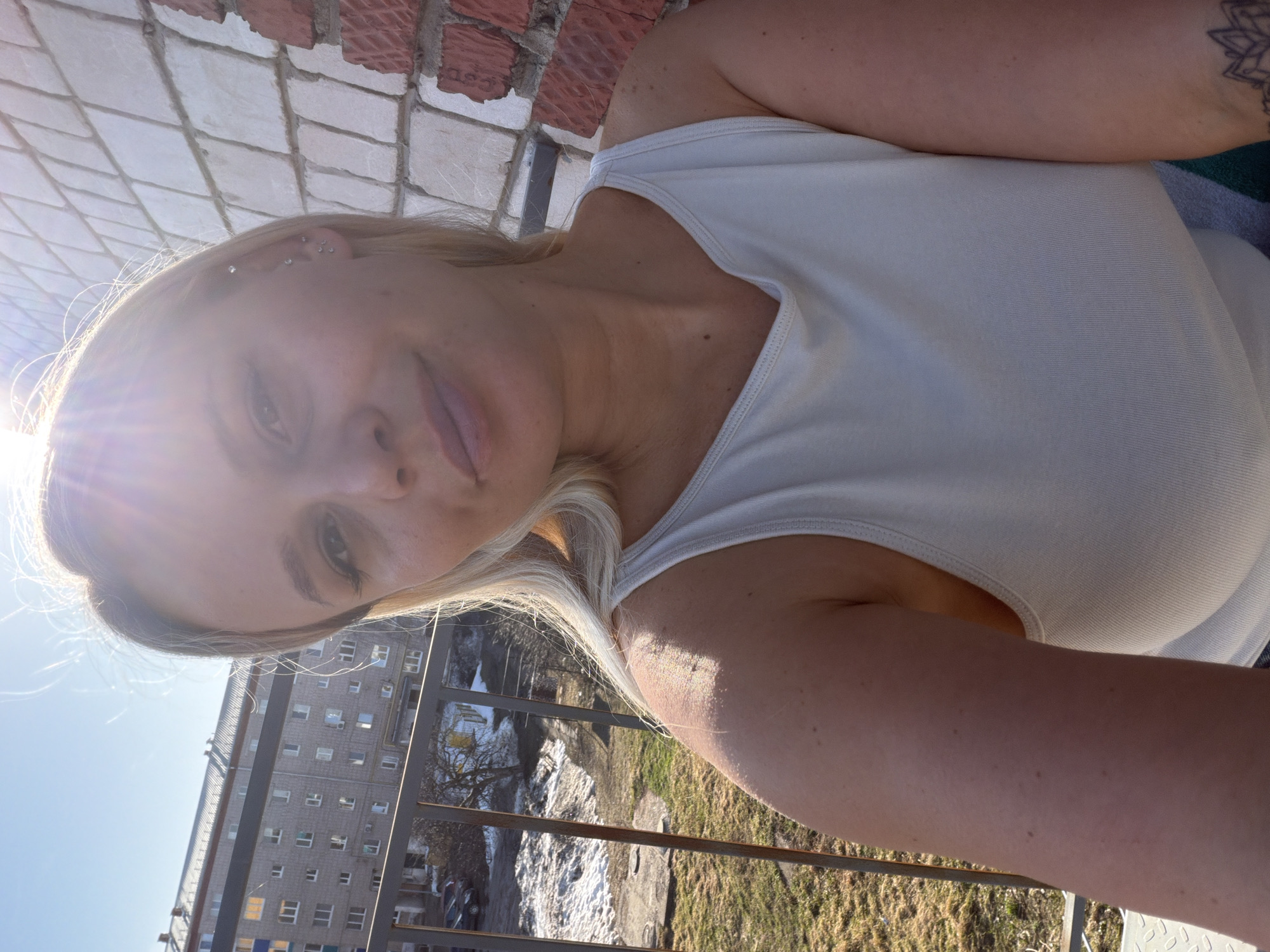 A medium shot, taken from a high angle, depicts a fair-skinned woman with blonde hair. She is wearing a white tank top and is positioned on a balcony.
The background reveals an urban landscape. A large, multi-story residential building is visible, and below the balcony, there is patchy snow in an open field.
The location is implied to be in the city of Sarapul, Russia.