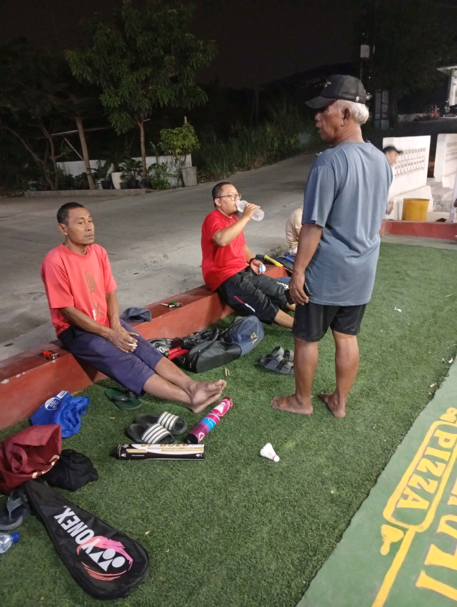 In the city of Caringin, Indonesia, three men are visible on a grass-covered surface adjacent to a red concrete border. One man, wearing a red t-shirt and blue pants, is seated on the red border, looking away from the camera. Another man, in a red t-shirt and black shorts, is seated next to him, drinking from a clear plastic bottle. A third man, wearing a blue t-shirt, black shorts, and a black baseball cap, stands facing the other two men. He has grey hair visible from under the cap. Several bags, a badminton racket in a case, flip-flops, a shuttlecock, and other unidentifiable objects are scattered on the grass. A portion of a yellow-lettered sign reading "PIZZA" is visible on the green surface. The lighting suggests it is nighttime.