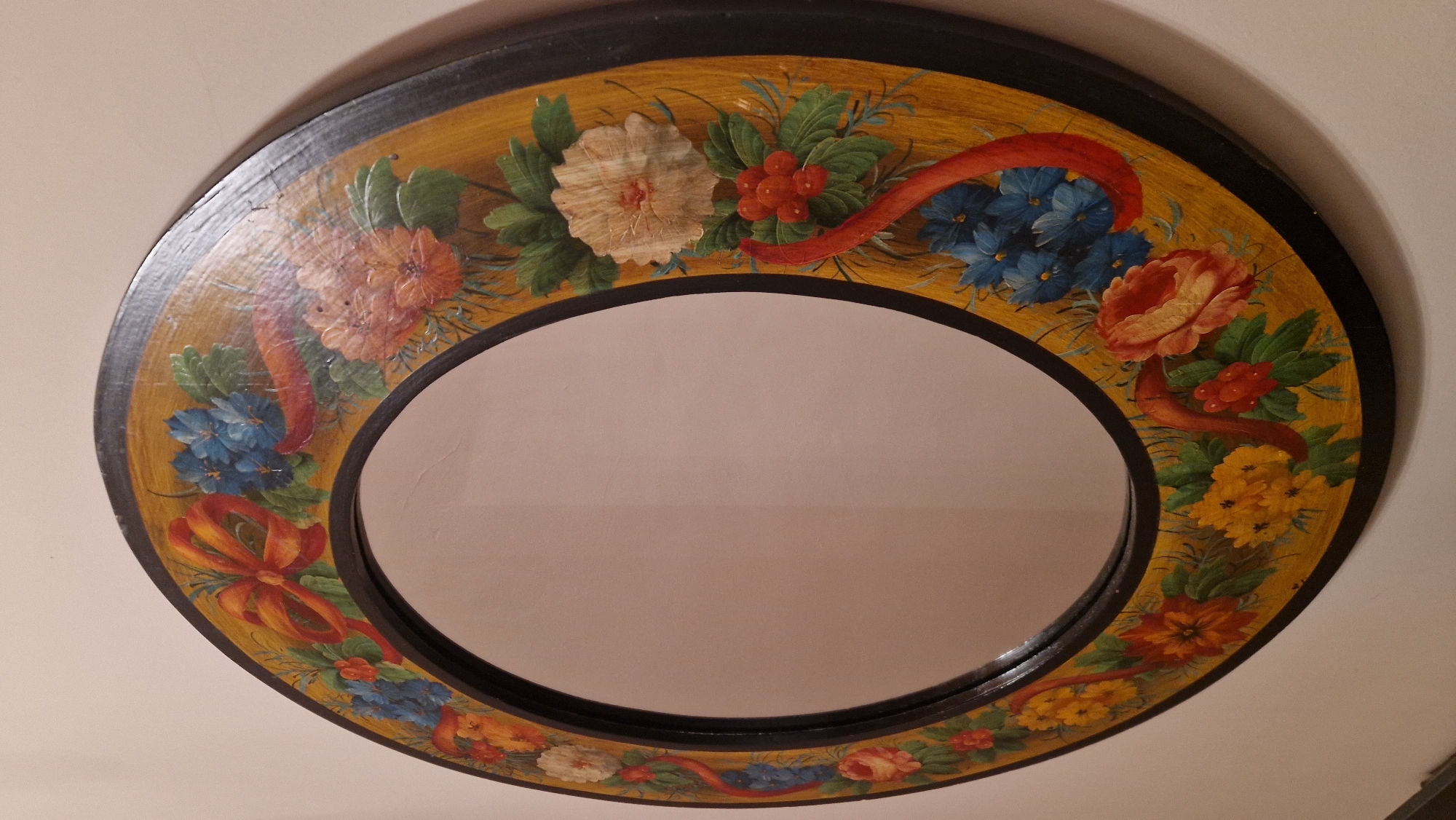 The image displays a round, decorative mirror with a hand-painted floral design around its frame. The frame itself is black, and the painted border is a golden yellow with various flowers and red ribbons depicted in an art nouveau style. The flowers are in shades of orange, red, yellow, and blue, with green leaves. The mirror is positioned on a light-colored wall, casting a subtle shadow. The image does not provide any information or visual cues that would allow for its location to be identified within the city of Vernate, Italy. There are no people or specific objects that would indicate a particular location.