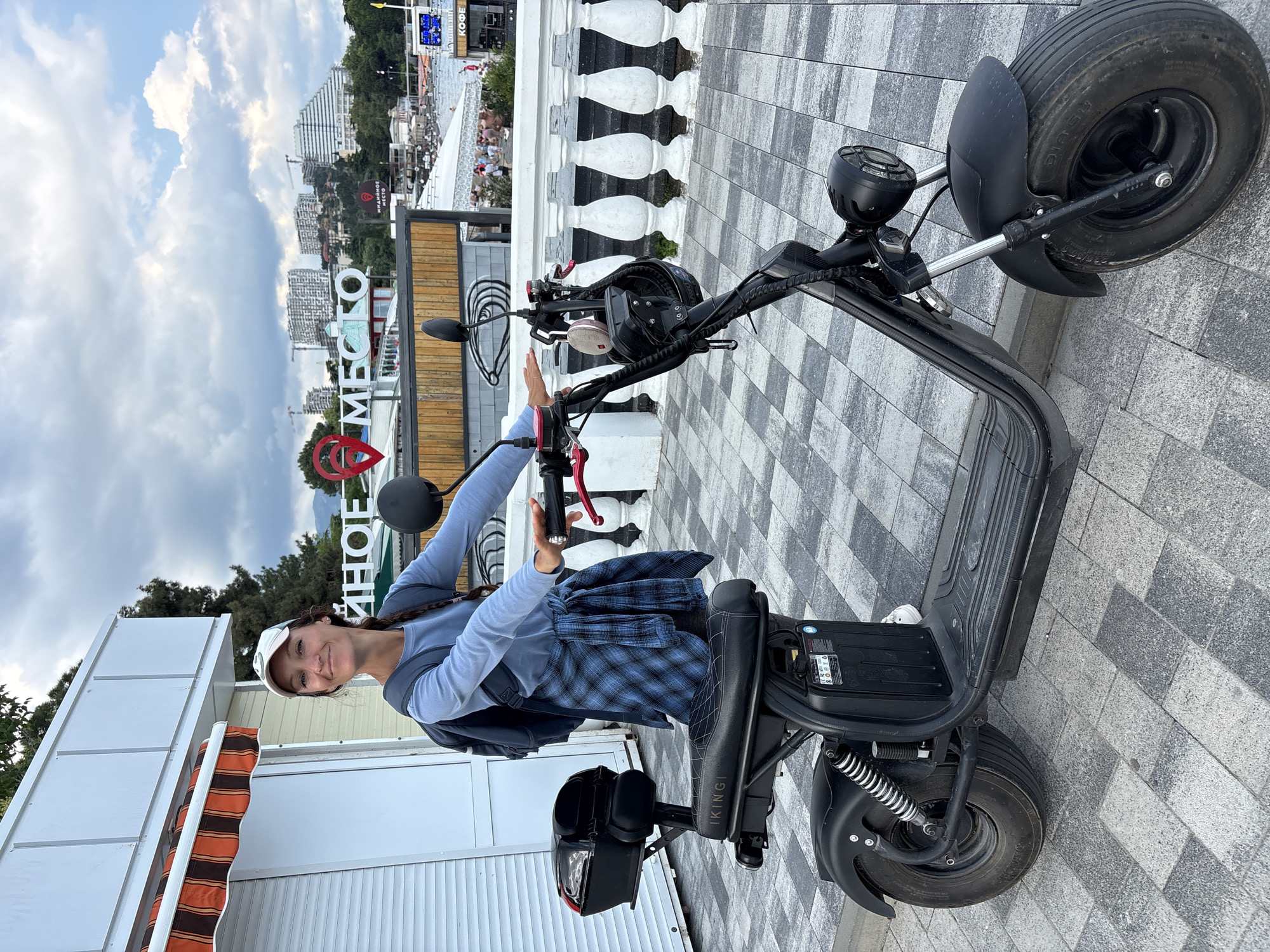 A woman is posing with a black electric scooter on a tiled outdoor walkway. She is wearing a blue long-sleeved shirt, a blue and white checkered shirt tied around her waist, a white baseball cap, and white sneakers. She has a backpack on. The scooter has a black seat, a storage compartment at the back, and a round headlight. In the background, there is a building with an orange and white striped awning, a white railing, and some city buildings and trees. The sky is cloudy. There is a sign in Russian that reads "ИННОЕ МЕСТО". The location is Alushta, Ukraine.