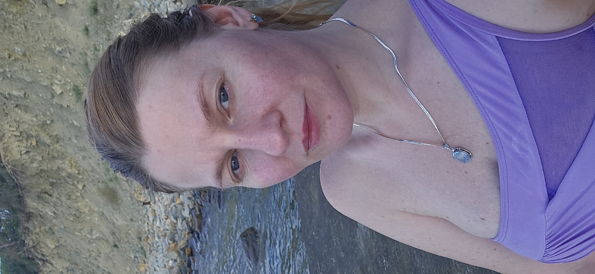 A medium shot, taken from a low angle, shows a fair-skinned woman with light brown hair pulled back into a ponytail. She is looking directly at the camera with a neutral expression. She has blue eyes and light makeup. She is wearing a silver chain with a light blue pendant and a lavender colored swimsuit. The background consists of a rocky shoreline and water. The scene is set in Dyurso, Russia.