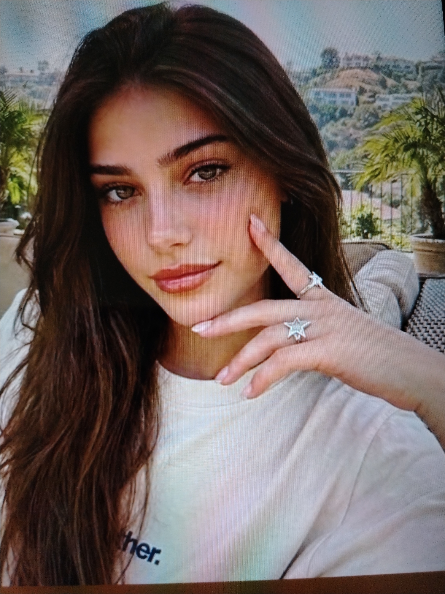 A young woman with long, dark brown hair and light brown eyes is shown from the chest up. She is wearing a light-colored t-shirt with the word "ther." visible on the front. She has a hand raised to her face, with two rings on her fingers. One ring is a silver band with a small star, and the other is a larger silver ring with a star design. She is looking towards the camera with a slight smile. The background appears to be an outdoor setting with palm trees and houses on a hillside, suggesting a warm climate. There is no indication that this image is located in Stavropol, Russia.