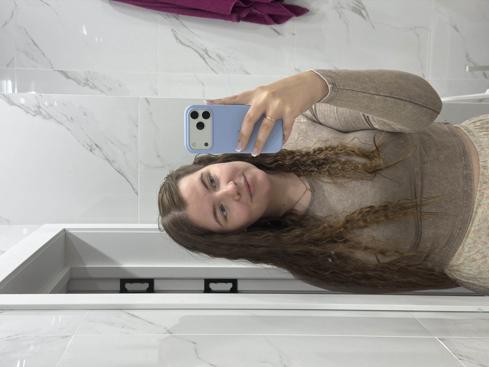 A young woman with light skin and curly brown hair is taking a selfie in a bathroom. She is wearing a long-sleeved beige top and a floral print skirt. She is holding a light blue phone with a triple-lens camera. She is wearing a gold ring on her ring finger. The bathroom has white marble tiles on the wall. A purple towel hangs in the background. There is a white countertop and sink in the foreground. This image was taken in the city of Chaikovsky, Russia.