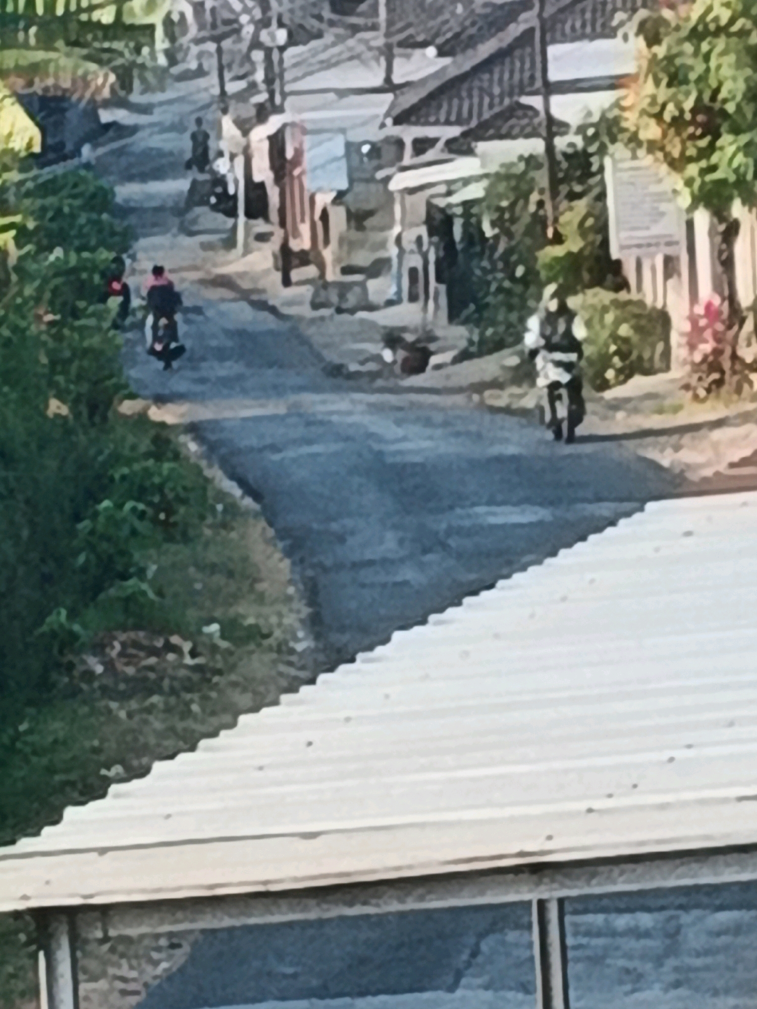 The image depicts a street scene in Bocek, Indonesia. A group of people on motorcycles are visible on an asphalt road that curves through a village. The road is flanked by houses with tiled roofs and lush green vegetation. The lighting suggests it is either morning or late afternoon. In the foreground, a corrugated metal roof is prominent.