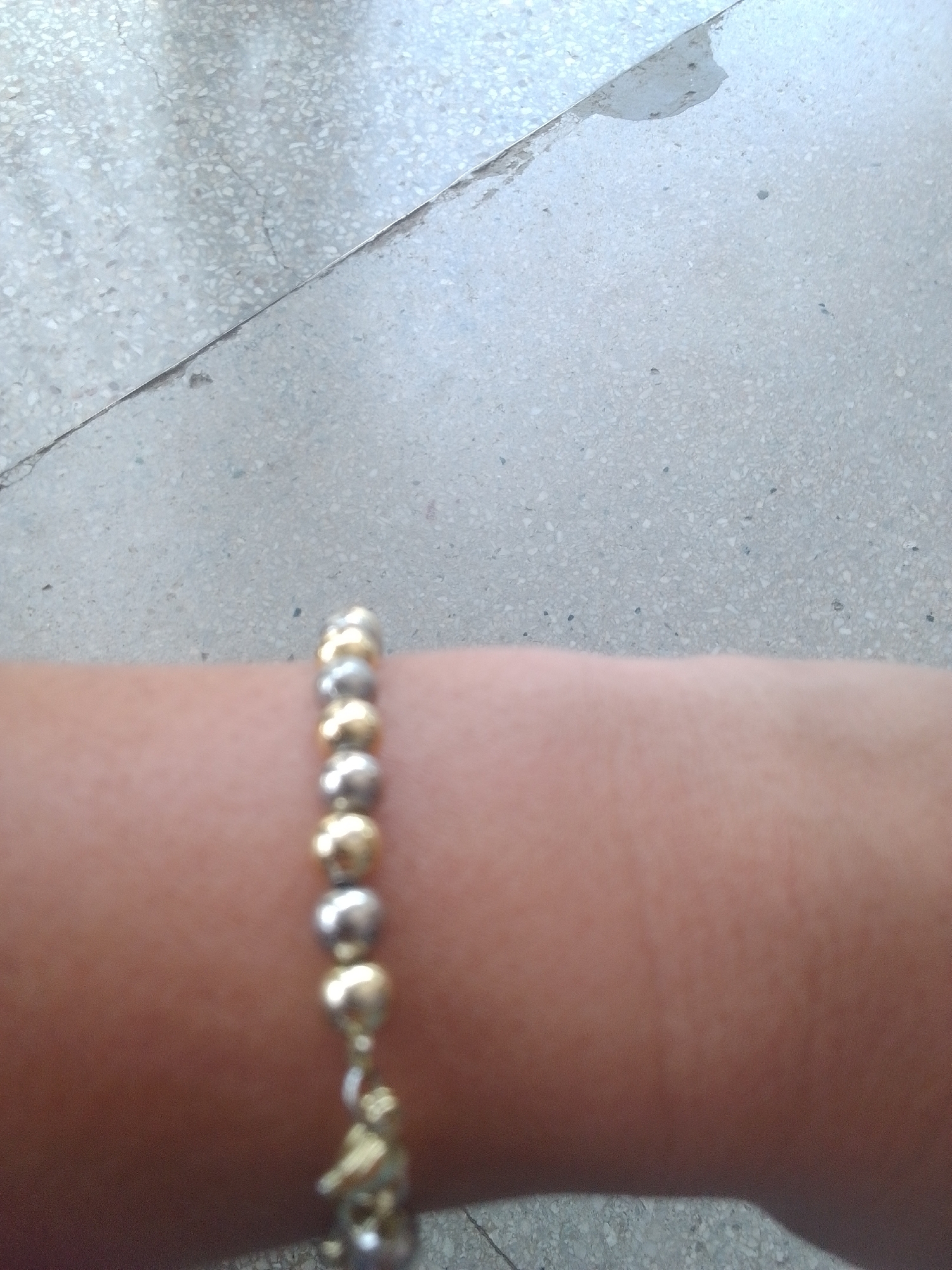 This is a close-up, horizontal, outdoor, eye-level shot focused on a bracelet worn on a person's arm. The bracelet is made of alternating gold and silver beads, with a small, gold charm hanging from it. The person's skin tone is visible as a warm, peachy color, and the texture of the skin is slightly blurred. The background consists of a textured, light-colored surface, possibly concrete or terrazzo flooring, with some subtle cracks and a slight sheen, suggesting it might be damp or recently cleaned. The lighting appears to be natural, casting soft shadows, and the overall ambiance is casual and everyday. There is no visible text in the image. The location context of La Tagua, Venezuela, does not provide specific visual cues within the image itself but suggests a tropical or subtropical climate, which could be consistent with the lighting and the casual nature of the scene. The activity depicted is simply the wearing of a piece of jewelry.