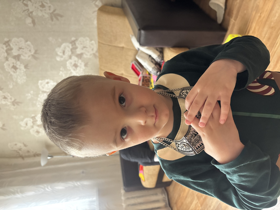 This image depicts a young child in an indoor residential setting in Pavlovsky, Russia. The child, with short light brown hair, dark eyes, and a neutral expression, faces directly towards the camera. He is wearing a long-sleeved dark green top featuring a cream-colored collar and a diagonal red and white striped emblem on the lower right front. A prominent silver-toned metal necklace, adorned with intricate geometric and stylized animal patterns on a shield-like central pendant, is worn around his neck. His hands are clasped together in front of his chest, resting over the necklace.
The background reveals a room with light-colored wallpaper displaying a repeating faint floral pattern. To the left, a window provides natural light, beneath which a white radiator is visible, and a floor lamp with a white shade stands nearby. A dark-colored sofa or armchair is partially visible on the left. To the right, a light brown sofa is present, upon which a red box, white fabric items, and other miscellaneous objects are placed. A dark, rectangular furniture item or object is partially visible on the far right. The floor is light-colored, appearing to be wood or laminate, and a white slipper is visible on the floor to the right.