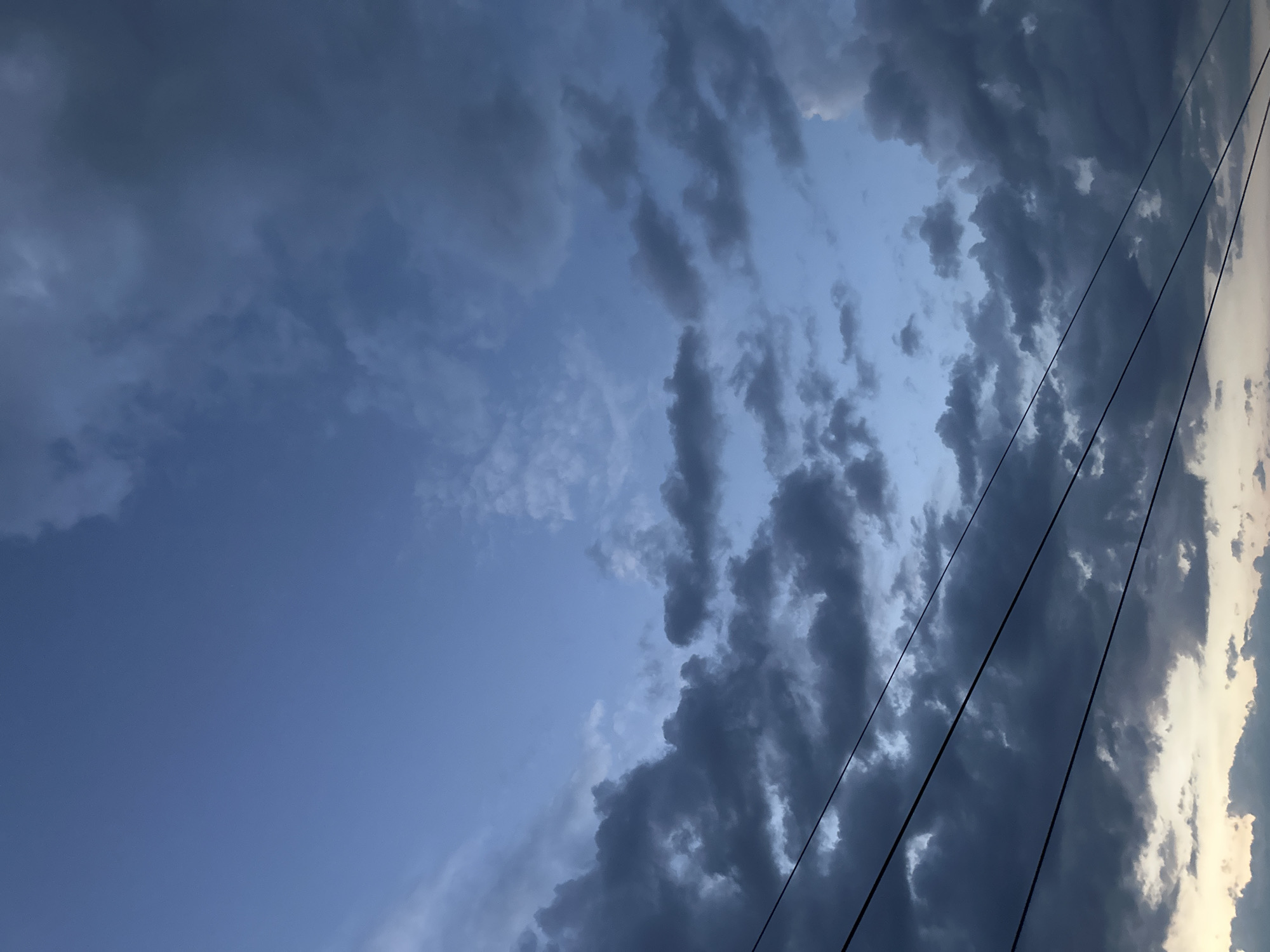 A low-angle shot captures a twilight sky filled with dramatic, dark clouds. Three thin, black power lines stretch diagonally across the lower portion of the frame. The sky transitions from a pale, dusky blue at the top to darker, more ominous grays and blues closer to the horizon. Some lighter clouds are visible against the darker masses. The image depicts an outdoor scene, likely in Energetik, Russia, during the evening. No people or specific structures are visible, with the focus solely on the sky and the power lines.