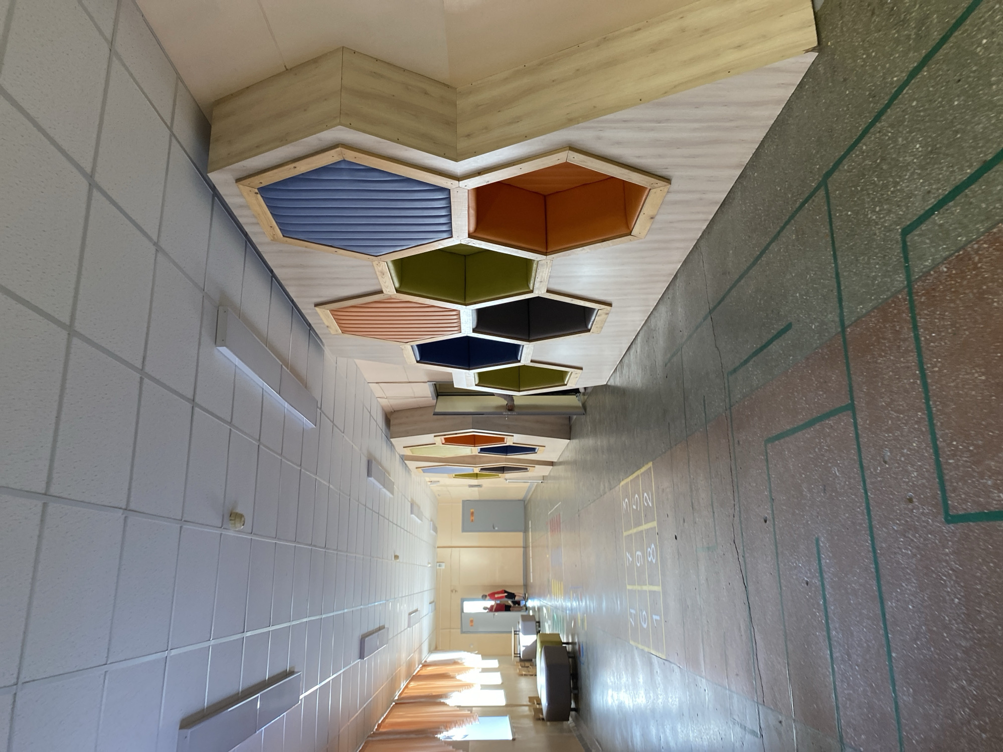 The image depicts a hallway interior, likely in a school or similar institutional building. The hallway features hexagonal ceiling decorations in alternating colors. The walls on the left side are white with evenly spaced rectangular light fixtures. The floor is composed of mottled brown and tan material, overlaid with green geometric patterns and yellow hopscotch squares. People are visible further down the hallway. The location is Votkinsk, Russia.