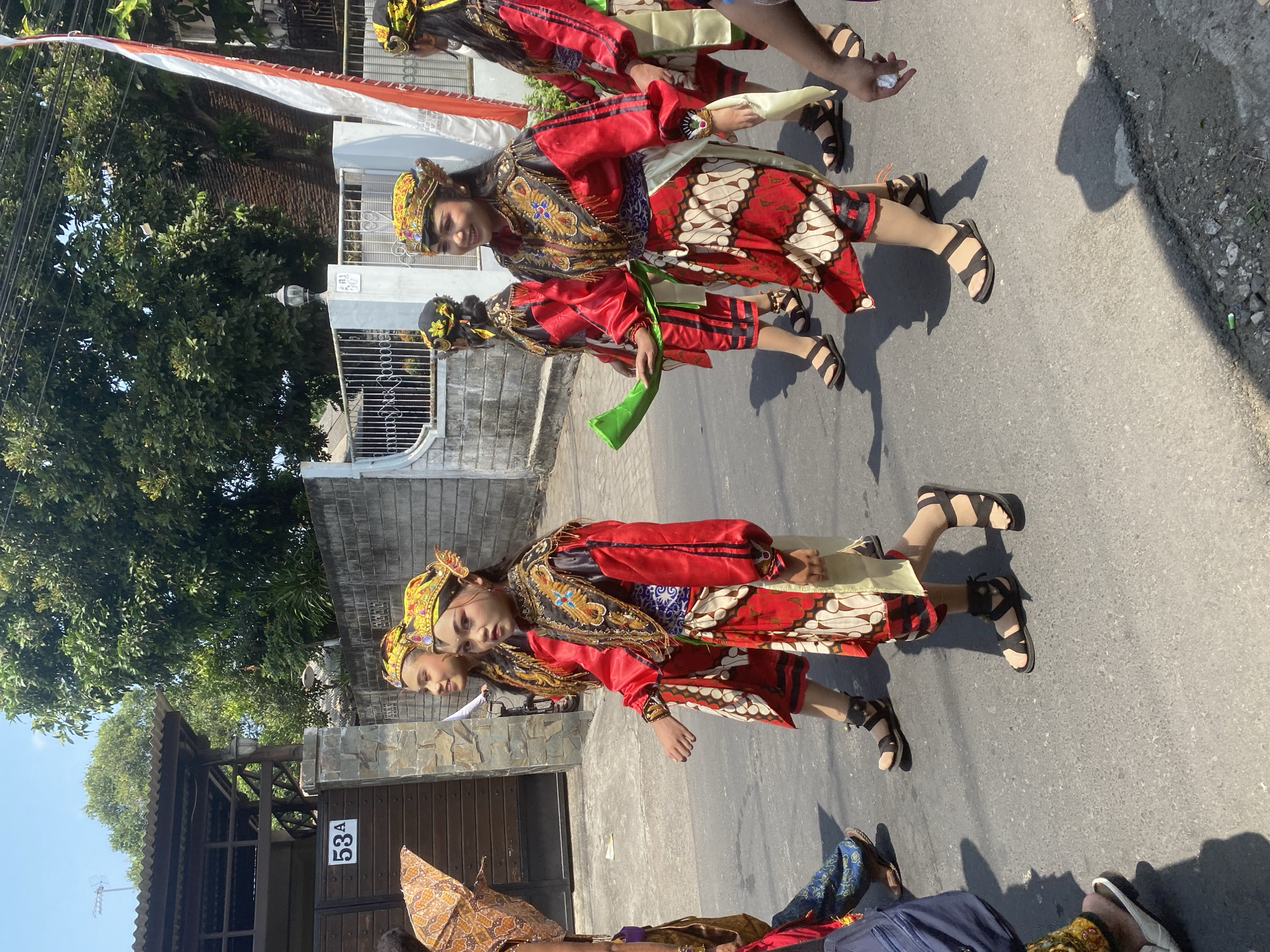In Kediri City, Indonesia, a group of individuals, likely performers, are walking down a paved street. The people are wearing traditional Indonesian attire, characterized by vibrant red tops with intricate gold and black embellishments, patterned skirts, and ornate headwear. Some are also wearing white sashes and dark sandals. One individual in the foreground is holding a green, horn-like object. The background features a stone wall, a gate, trees, and a building with a visible house number "53A". A red and white Indonesian flag is also present. The scene appears to be a daytime outdoor event or procession.