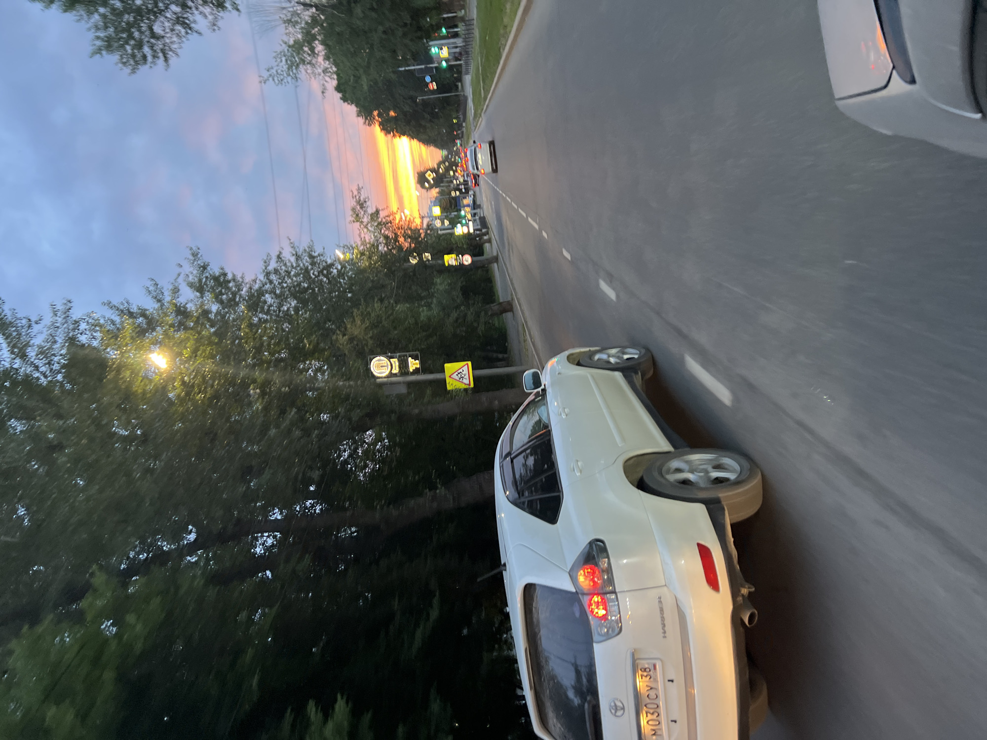 The image depicts a road scene in Irkutsk, Russia, during a sunset. The sky is filled with dramatic colors, ranging from soft pinks and oranges to deep blues, indicating the end of the day. Tall, dark green trees line the left side of the road, their leaves illuminated by streetlights.
The primary subject is a white SUV, seen from the rear-right perspective, in motion on the asphalt road. The car has its taillights illuminated, suggesting it is either driving or has recently stopped. Its license plate is visible and reads "M030CY 38". Other vehicles are present further down the road, creating a traffic flow.
Several traffic signs are visible along the roadside. One sign, a yellow diamond with a black border and a black pedestrian symbol, indicates a pedestrian crossing. Another circular sign with a yellow background and a black border features a black "T" shape, possibly denoting a specific road or traffic regulation. Streetlights are also scattered along the road, some casting pools of light on the trees and the road surface.
The overall mood is one of a peaceful evening commute or journey, enhanced by the vibrant sunset colors. The motion blur in the image suggests the photo was taken from another moving vehicle. The presence of Russian license plates and Cyrillic text on the signs firmly places the location in Russia.
