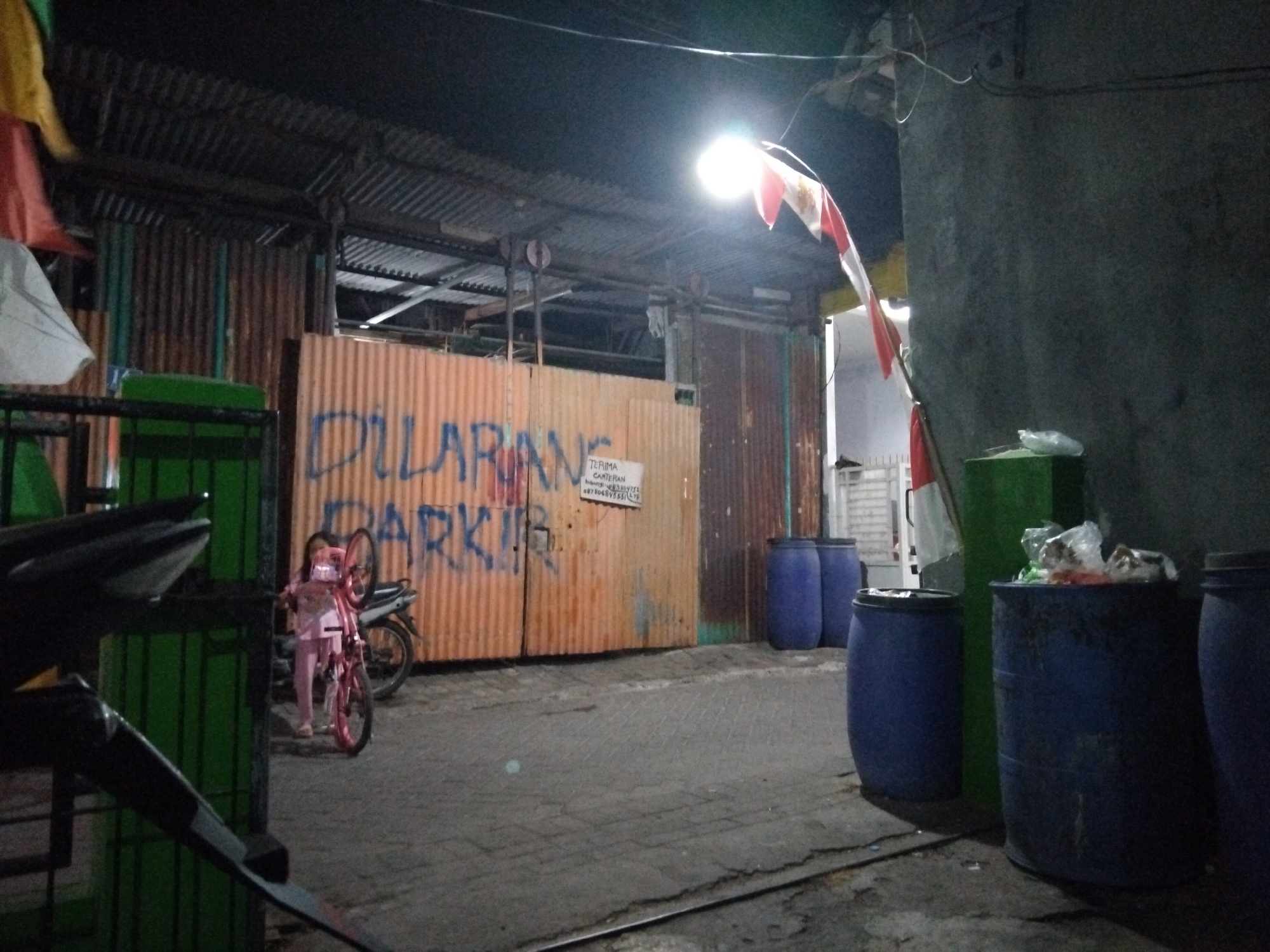 In the city of Kepuh Kiriman, Indonesia, a nighttime scene depicts an alleyway with a corrugated metal fence. A young girl, dressed in pink clothing, stands beside a pink bicycle. The fence has large blue lettering that reads "DILARANG PARKIR," meaning "NO PARKING." To the right of the fence, three large blue barrels are visible, one of which is filled with trash. A bright overhead light illuminates the area. The ground is paved with cobblestones and a metal track runs through it.