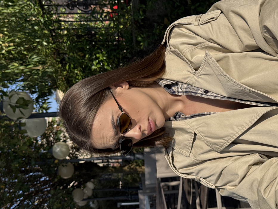 The image portrays a young adult female, captured from the mid-chest upwards, situated in an outdoor public area within Sirmione, Italy. She wears dark, oval-lensed sunglasses with thin metal frames, an unbuttoned light-colored plaid shirt, and an unbuttoned beige trench coat featuring lapels and shoulder epaulets. Her medium-long brown hair is parted on the left. The subject's gaze is directed downward and slightly to the left of the frame, exhibiting a neutral expression. The background is blurred, showing dense green foliage on the right, dark outdoor tables and chairs on the left, and several street lamps with spherical frosted globes positioned in the upper left portion of the scene. Strong sunlight illuminates the environment, creating distinct highlights and shadows.