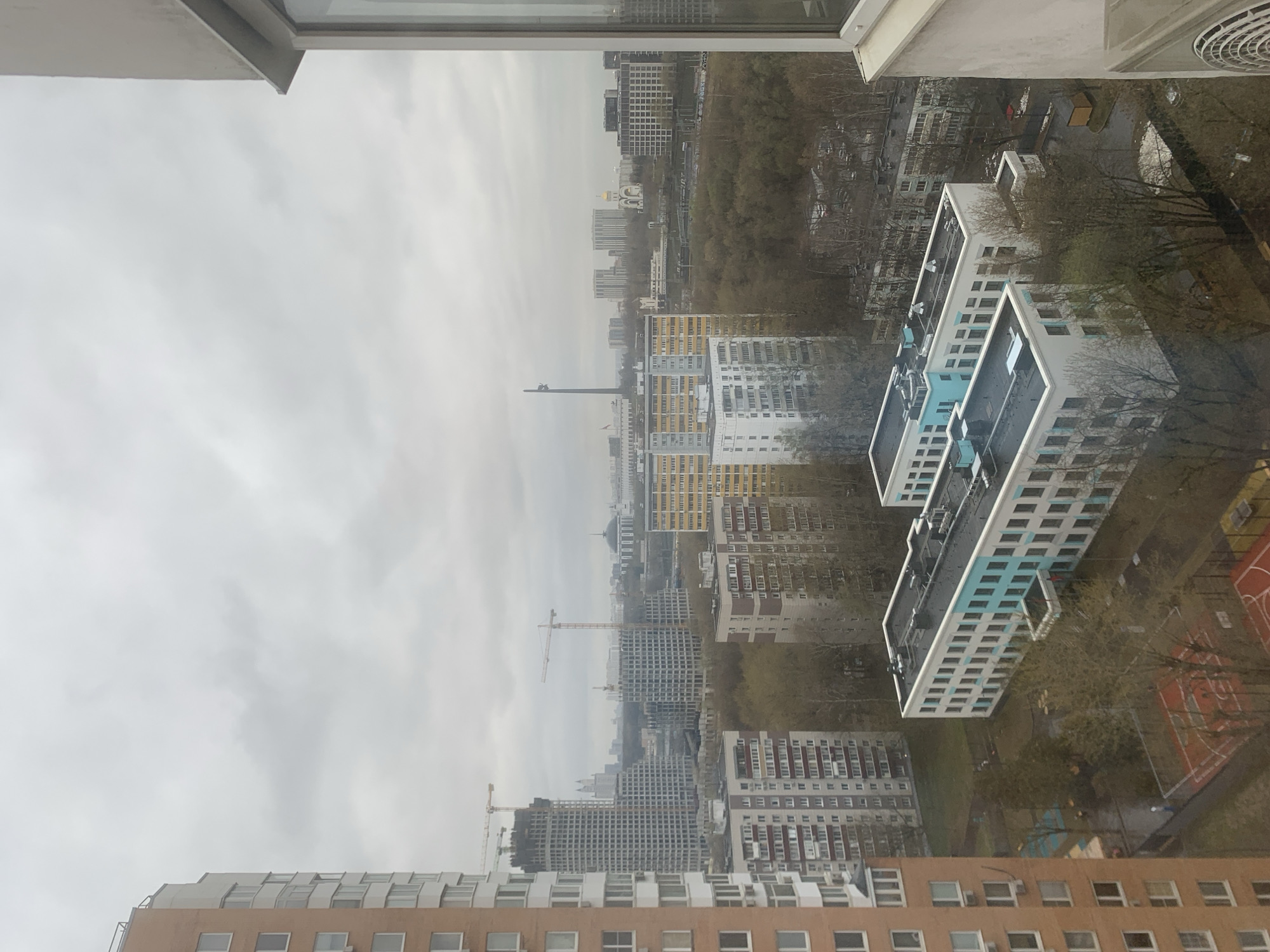 This is a photograph taken from an elevated position in Moscow, Russia, looking out over a dense urban landscape on an overcast day. The sky is uniformly gray. A mix of mid-rise and high-rise residential buildings dominate the view, showing a variety of architectural styles and colors. There are some buildings undergoing construction, evidenced by the presence of cranes. Some buildings have facades in white and blue, while others are yellow. Interspersed with the buildings are trees and patches of green space, suggesting parks or courtyards. In the foreground, the edge of the window frame and what appears to be a part of an air conditioning unit are visible. A basketball court is visible in the bottom right. The scene suggests a residential area with ongoing development.
