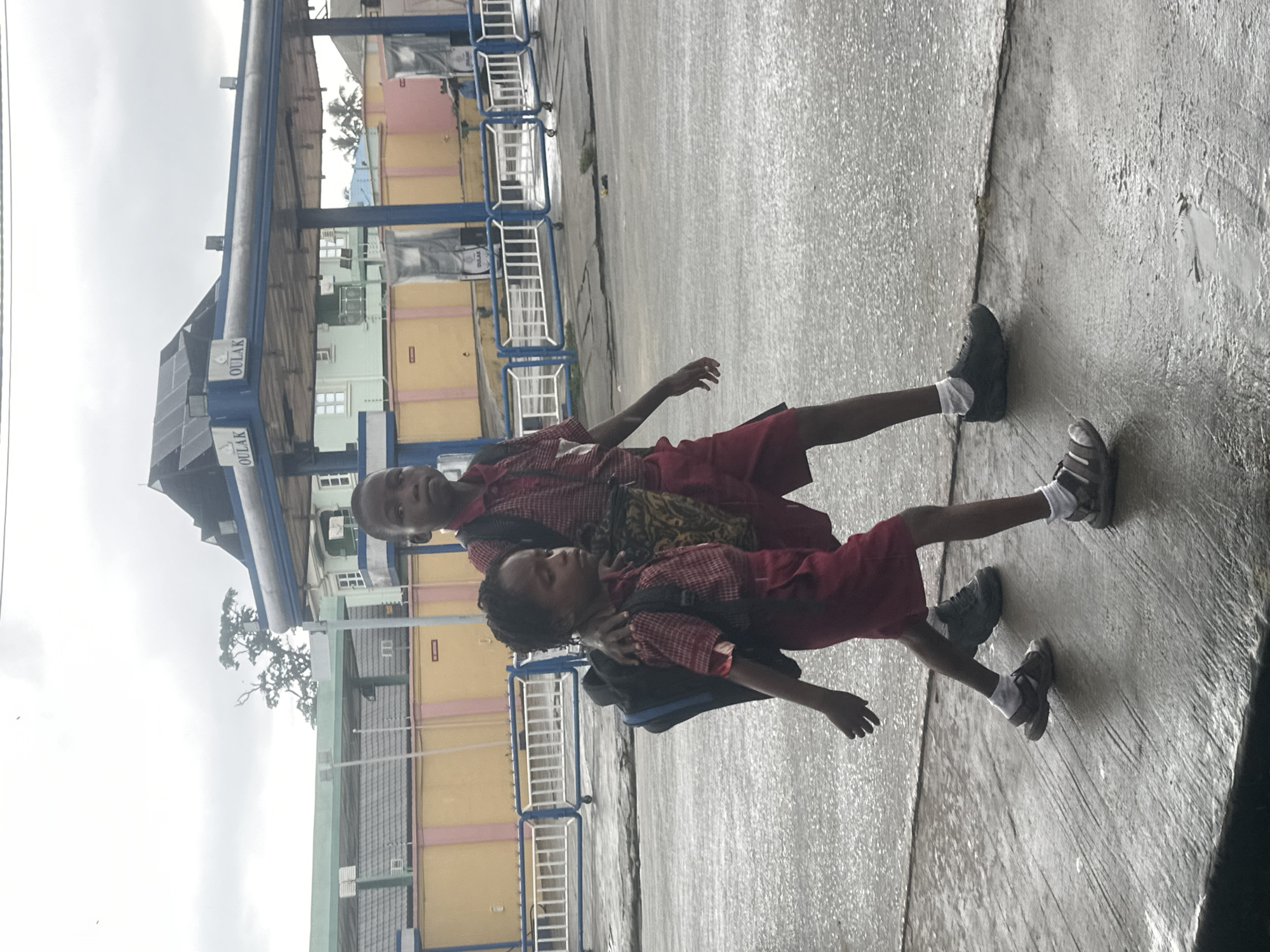 Two young children, likely siblings, are walking on a wet asphalt street in front of a covered structure with "OULAK" signage. The taller child, a boy, is walking slightly ahead and to the right of the younger child, a girl. Both are wearing school uniforms consisting of red plaid shirts and red shorts or skirts. The boy has a black backpack, while the girl also carries a black backpack and a patterned bag slung over her shoulder. The boy has his arm around the girl's shoulder in a protective gesture. They are walking away from the camera towards the left of the frame. The ground is wet and reflective, indicating recent rain. In the background, there are buildings and what appears to be a gas station canopy. The sky is overcast. The scene is set in the city of Badagry, Nigeria.