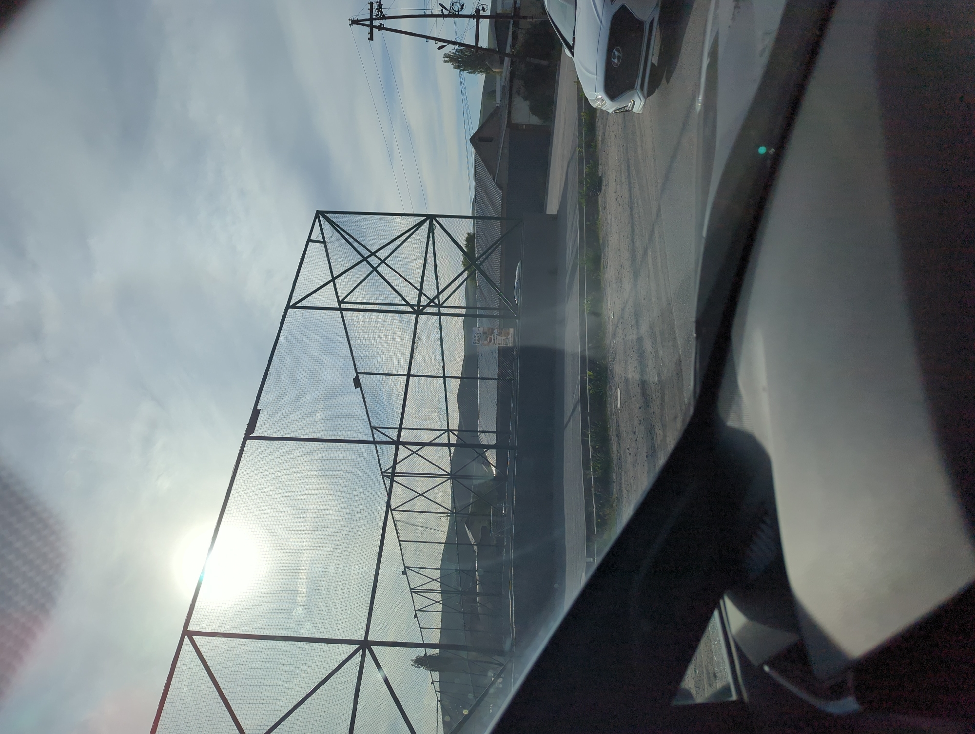 A ground-level, eye-level, medium-shot photograph taken from the interior of a vehicle shows a wire fence in Beş-Küŋgöy, Kyrgyzstan. The fence is made of metal posts and wire mesh and extends along the left side of the frame. The sun shines brightly through the fence mesh. A street and a parked white vehicle are visible behind the fence, along the right side of the frame. Overhead power lines are visible against the sky. Part of the interior of the vehicle fills the right foreground. The sky is partly cloudy.