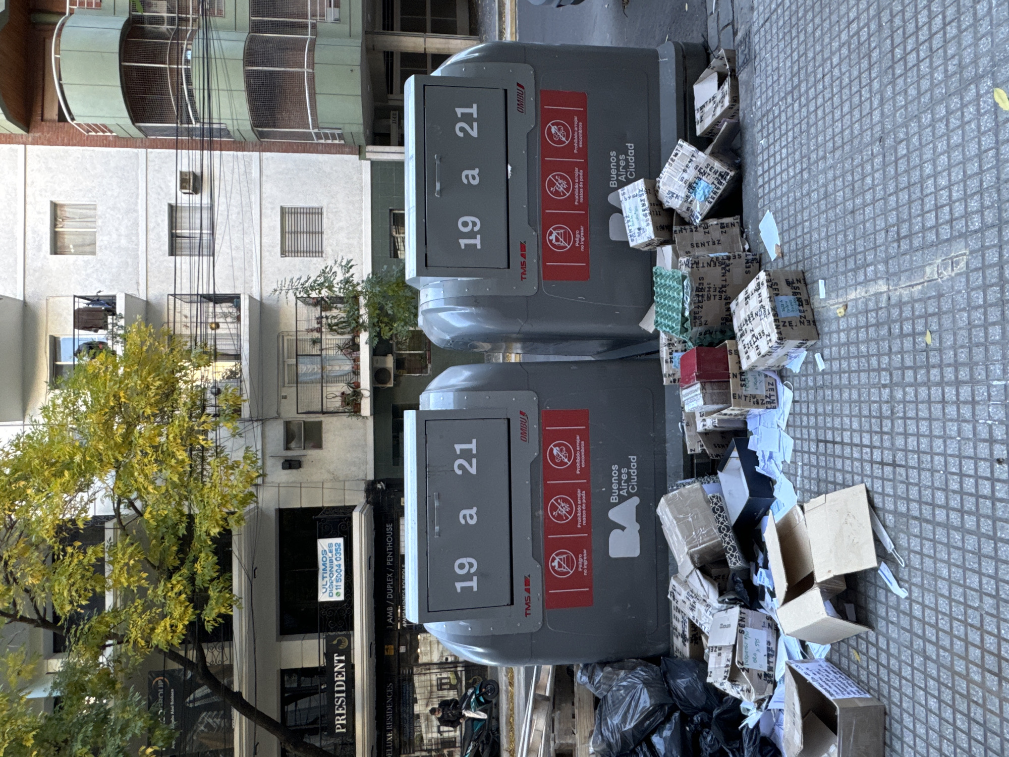 The image depicts a street scene in Buenos Aires, Argentina. Two gray, above-ground dumpsters are positioned on a tiled sidewalk. Each dumpster features signage indicating the permissible disposal hours (19 to 21) and warnings against throwing debris or garden waste. The "Buenos Aires Ciudad" logo is visible on the side of the dumpsters.
Scattered around the base of the dumpsters are numerous discarded cardboard boxes, some of which are open and revealing contents. Additional refuse, including black plastic bags, is piled at the base of the containers.
In the background, buildings of varying heights and architectural styles line the street. A tree with yellowing leaves is situated to the left, partially obscuring a building with signage that reads "PRESIDENT" and "DELUXE RESIDENCES." The sign also indicates the availability of apartments, duplexes, and penthouses, along with a phone number.