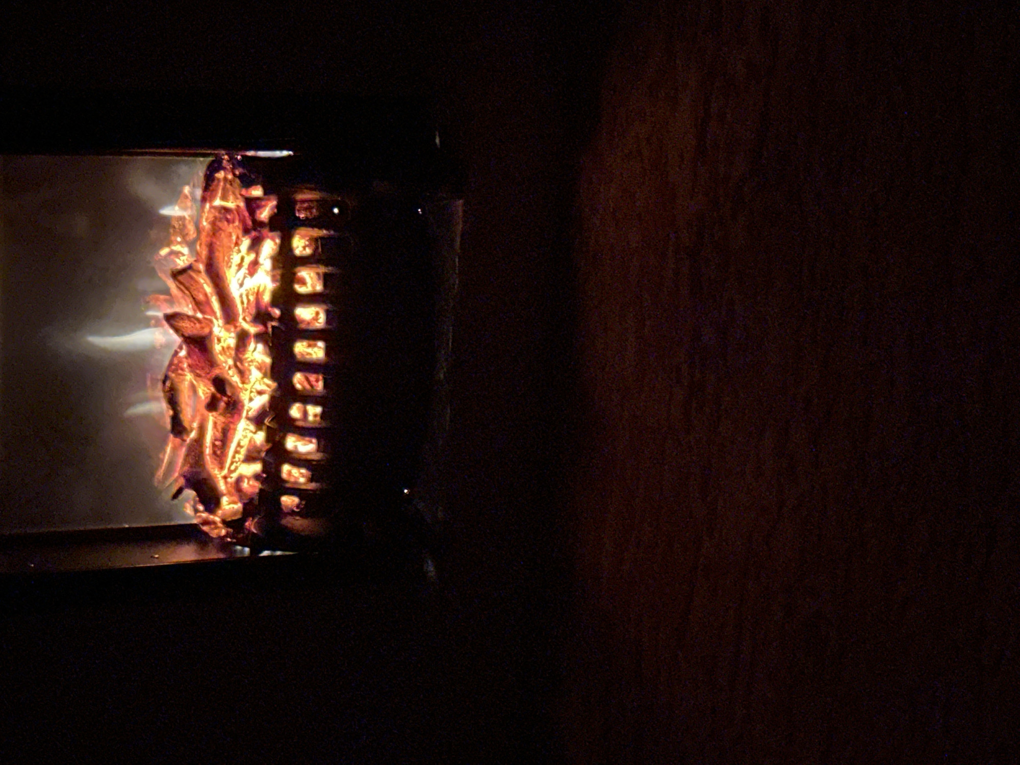 The image shows a close-up of an electric fireplace with glowing faux logs in a dark room. The logs are arranged in a metal grate, emitting a warm, orange light. Wisps of smoke-like steam rise from the logs, adding to the illusion of a real fire. The background is mostly dark, with the exception of the fireplace. The floor in front of the fireplace is a dark, textured surface, possibly wood or carpet. There are no people or animals visible in the image. The scene appears to be set indoors, likely in a living room or den. The lighting is dim, with the primary light source being the electric fireplace. The overall mood is cozy and atmospheric.