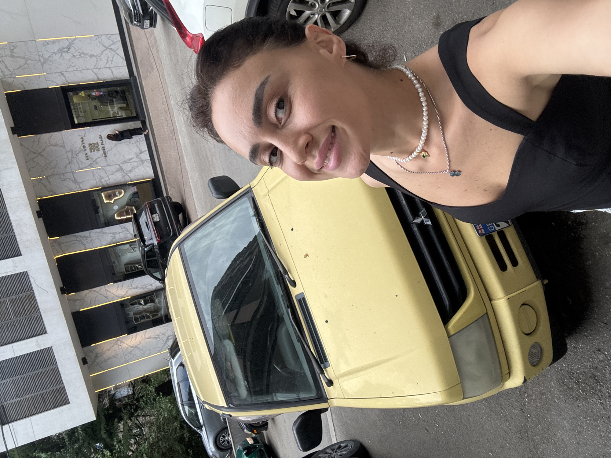 A woman is taking a selfie outdoors in front of a yellow vehicle, with a modern building in the background. The building features large windows, marble accents, and vertical yellow lighting. A sign on the building reads "SHOES PLAZA". Other vehicles, including a dark SUV and a silver car, are parked nearby on a paved surface. The woman is wearing a black top and a pearl necklace. The overall setting suggests an urban environment in Tbilisi, Georgia.