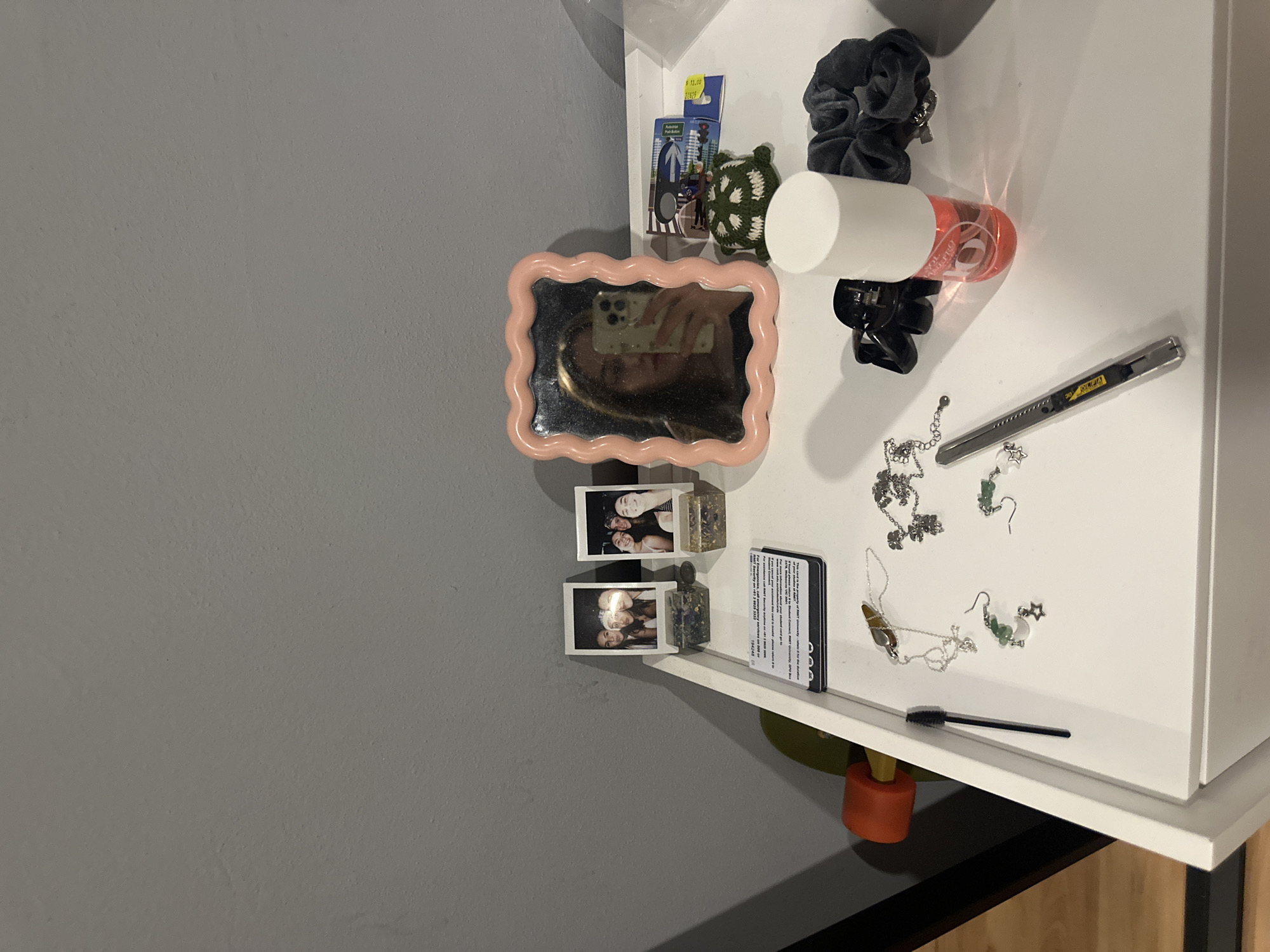 This is a medium shot of a shelf mounted on a wall in what appears to be an indoor setting, likely a bedroom or personal space. The wall is painted in a neutral gray tone, and the shelf is white. The items on the shelf appear to be arranged haphazardly.
Objects on the shelf include a pink, wavy-edged mirror reflecting a phone, next to the mirror is a small package, possibly a toy or trinket, a green crocheted item, a gray scrunchie, and a white and orange cylindrical object. There's also a black hair clip.
Scattered across the white surface of the shelf are various pieces of jewelry, including necklaces and earrings. A silver utility knife is placed near the jewelry. Two small framed photos and a business card or similar document are located on the left side of the shelf, closest to the wall. A black mascara wand is lying at the front edge of the shelf. The back half of a skateboard is visible underneath the shelf.
The scene provides a glimpse into the personal items of the space's occupant, suggesting a casual and lived-in environment. The location in Huixquilucan de Degollado, Mexico, cannot be verified from the image alone.