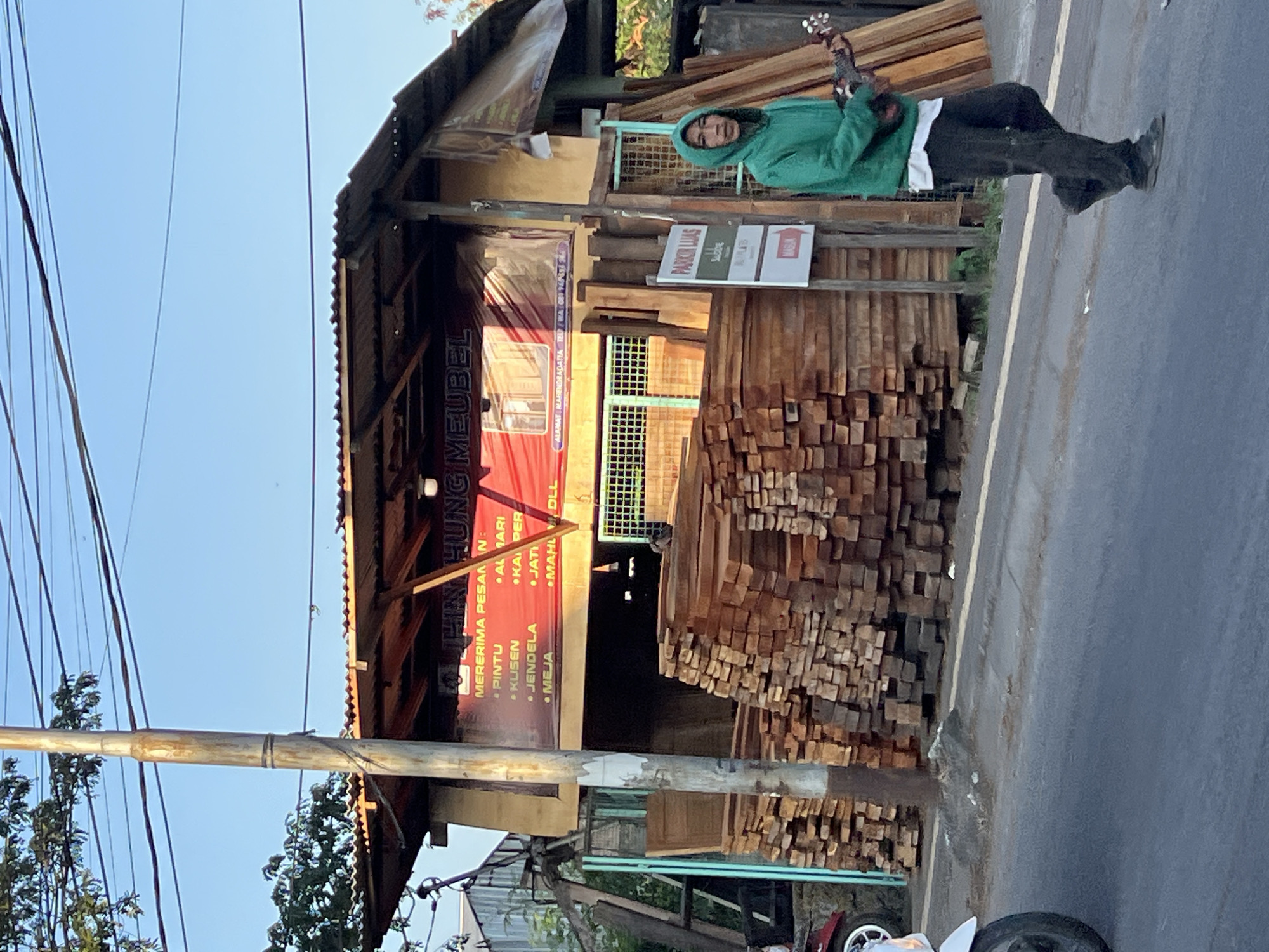 The image shows a street scene in Denpasar, Indonesia. In the background, a small wooden structure with a corrugated metal roof serves as a furniture shop. A red banner with Indonesian text, "TINH HUNG MEUBEL", is displayed on the front of the shop. Piles of lumber are stacked in front of and to the side of the shop. A person wearing a green hoodie and black pants is walking in the foreground, holding a ukulele. A signpost with the text "PARKIR LUAS" (meaning "ample parking") is visible to the left of the person. The street is paved, and a white line marks the edge of the road. Trees and power lines are also present in the scene. The sky is clear and blue, suggesting it is daytime.