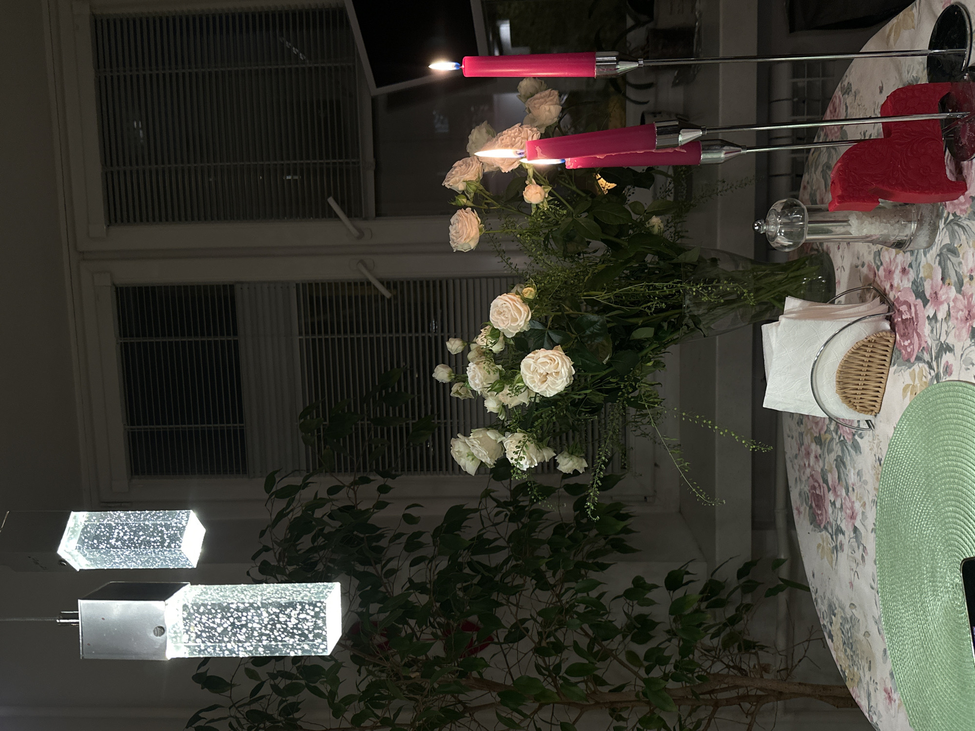 The media file displays an indoor scene, likely a dining or living area, during the evening or night. The primary focus is a round table adorned with a floral tablecloth featuring light-colored fabric with pink and green flower patterns. A mint green, circular woven placemat is prominently visible on the right side of the table.
A central element on the table is a glass vase holding a bouquet of white roses and various green leaves. Behind the roses, two slender, hot pink candles in silver-toned metal holders are lit, their small blue flames casting a warm glow. Nearby, a glass salt or pepper grinder, a small wicker basket, and a holder with white paper napkins are arranged on the table. A red decorative object, possibly a napkin holder, is also present next to the candles.
In the left foreground, two modern hanging light fixtures are visible. Each fixture consists of a clear, cube-shaped glass piece embedded with numerous small bubbles, emitting a soft white light. Behind these fixtures and partially obscured by them, a large potted plant with dark green foliage adds a natural element to the room. In the background, a window with white frames and horizontal blinds or shutters appears dark, indicating the exterior is dark.
The scene conveys a peaceful and intimate ambiance, suggesting a special occasion or a quiet dinner setting. No people are visible, and there is no visible text. The lighting primarily comes from the lit candles and the decorative fixtures.