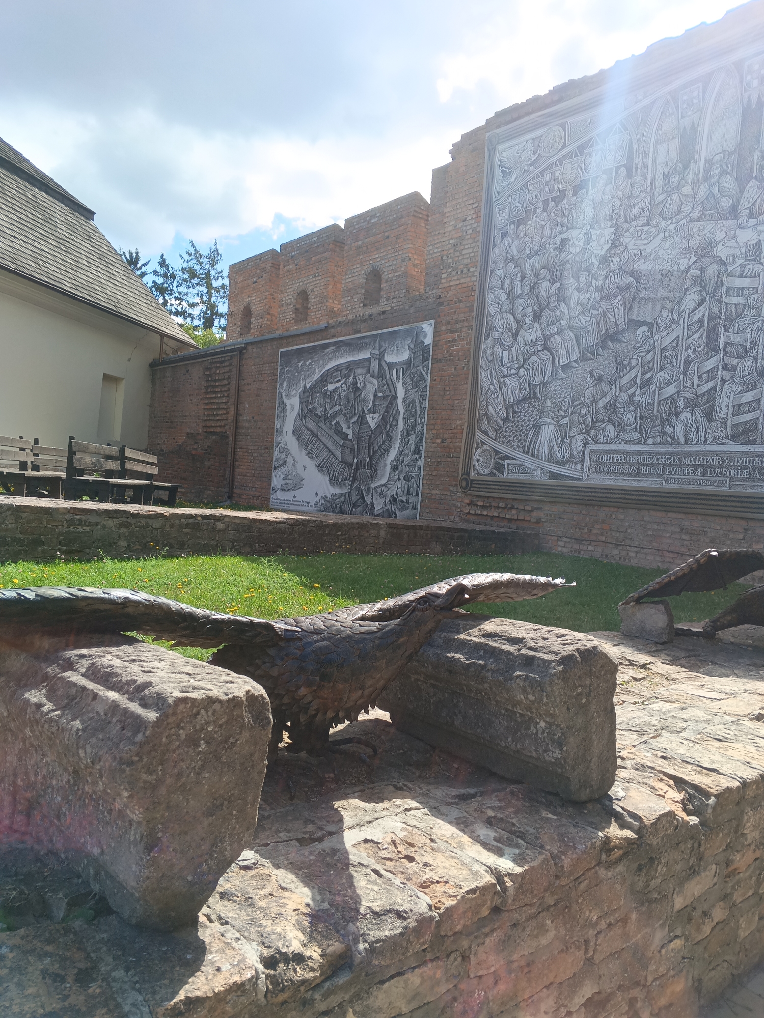 The image depicts an outdoor courtyard with several notable features. In the foreground, a stone sculpture of a bird, possibly an eagle, is positioned on a low brick wall. The bird sculpture is detailed and appears to be made of metal or a similar material. To the left, a building with a grey tiled roof and white walls is visible, with two wooden benches in front of it. Behind the benches and the bird sculpture, a brick wall serves as a backdrop. Mounted on this brick wall are two large, detailed black and white murals. The mural on the right is significantly larger and depicts a crowded scene with many figures, likely a historical event or assembly, with Latin text at the bottom. The mural on the left is smaller and appears to be a city or fortress plan. The environment suggests a historical or cultural site. This scene is located in the city of Lutsk, Ukraine, at the Lubart's Castle complex, specifically in the courtyard area. The sky above is blue with scattered clouds, and sunlight is casting shadows across the scene, indicating it is daytime. There are no people actively engaged in any activity within the frame.