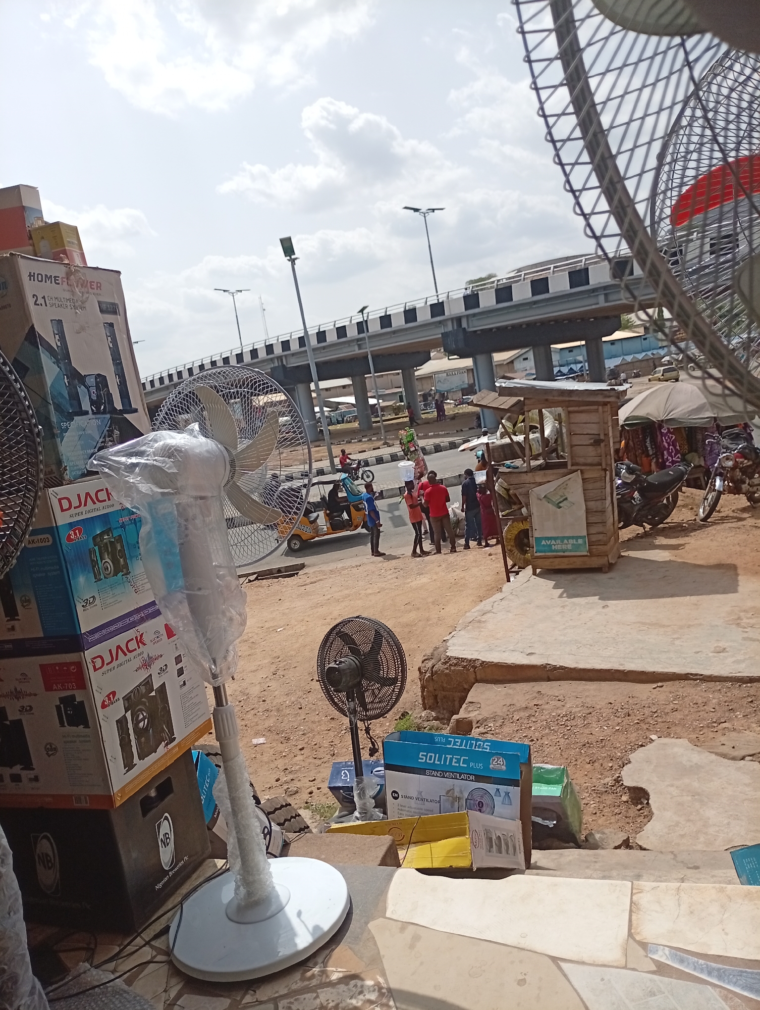 The image presents a street scene in Lokoja, Nigeria. An elevated highway structure looms in the background. In the foreground, an assortment of products for sale is visible, including several stand fans and boxes of "D JACK" audio equipment. People are gathered on the side of the road, including a few wearing red shirts. A yellow "keke" auto-rickshaw and motorcycles are also present. A small wooden kiosk with "AVAILABLE HERE" printed on it is next to a small market stall with an umbrella.