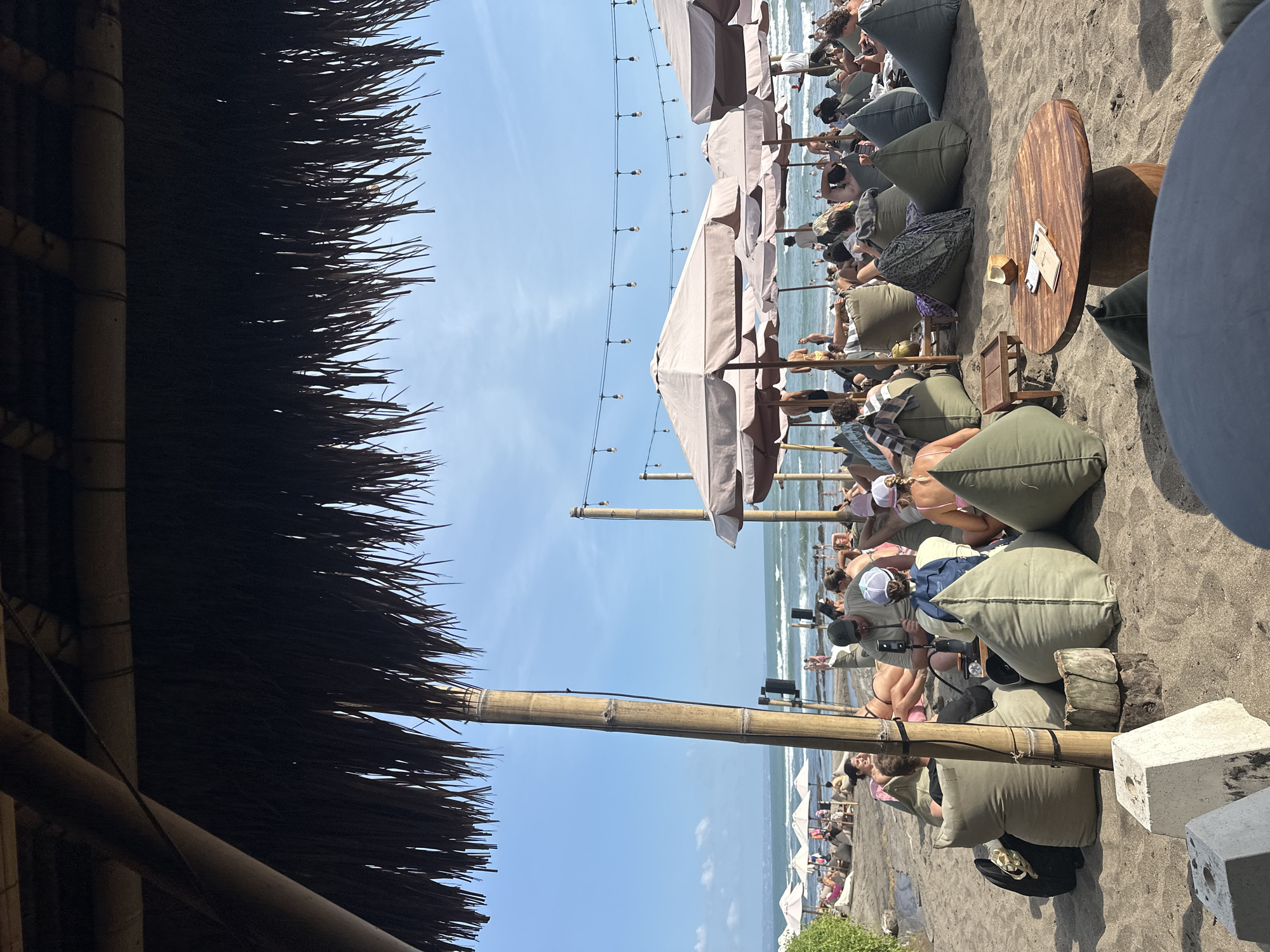 A group of people are relaxing on a beach in North Kuta, Indonesia. They are sitting on bean bag chairs and lounging under umbrellas. The ocean is in the background, with waves gently rolling onto the shore. The sky is blue with a few scattered clouds. String lights are hung between poles, adding to the ambiance of the beachside establishment. A table in the foreground holds a coconut and some menus.