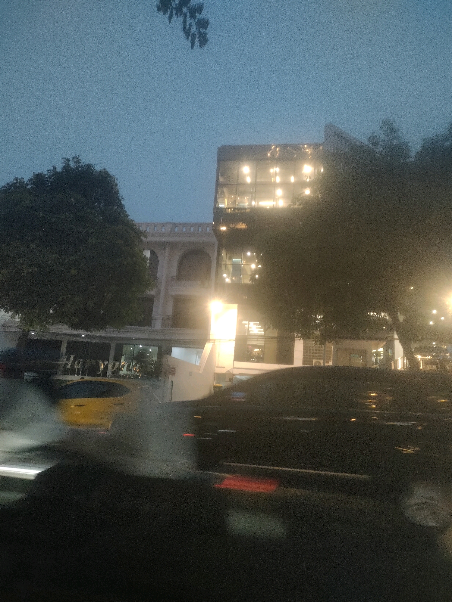 The image captures a street view in Surabaya, Indonesia, at dusk or night. The scene is dominated by a building with a modern glass facade, illuminated by interior lights, and an adjacent older building with arched windows and a decorative upper railing. In the foreground, several cars are visible, including a yellow car and two dark-colored vehicles. The street appears wet, suggesting recent rain or cleaning. Trees are present, partially obscuring the buildings. The overall impression is one of urban life transitioning into evening.