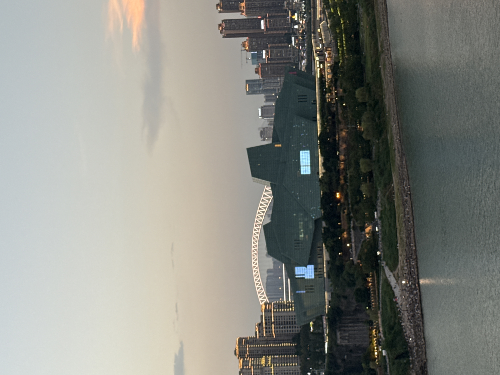 This media file depicts a city skyline at dusk, with a large body of water in the foreground. The scene is dominated by a cluster of modern skyscrapers, some illuminated with lights. A prominent green, angular building with blue-lit windows stands out among them. Behind this structure, a large, white arched structure, possibly a Ferris wheel or bridge, is visible and illuminated.
To the right of the buildings, a tree-lined park runs along the edge of the water, with pathways and scattered lights indicating human activity. A multitude of people are gathered along the waterfront, suggesting a public gathering or promenade. The water appears calm, reflecting the ambient light.
The sky shows the fading light of sunset, with some wispy clouds tinged with orange and yellow. The overall atmosphere is serene and urban, with a sense of a city winding down for the evening. The location is identified as Yuzhong District, China. No discernible text is visible in the image.