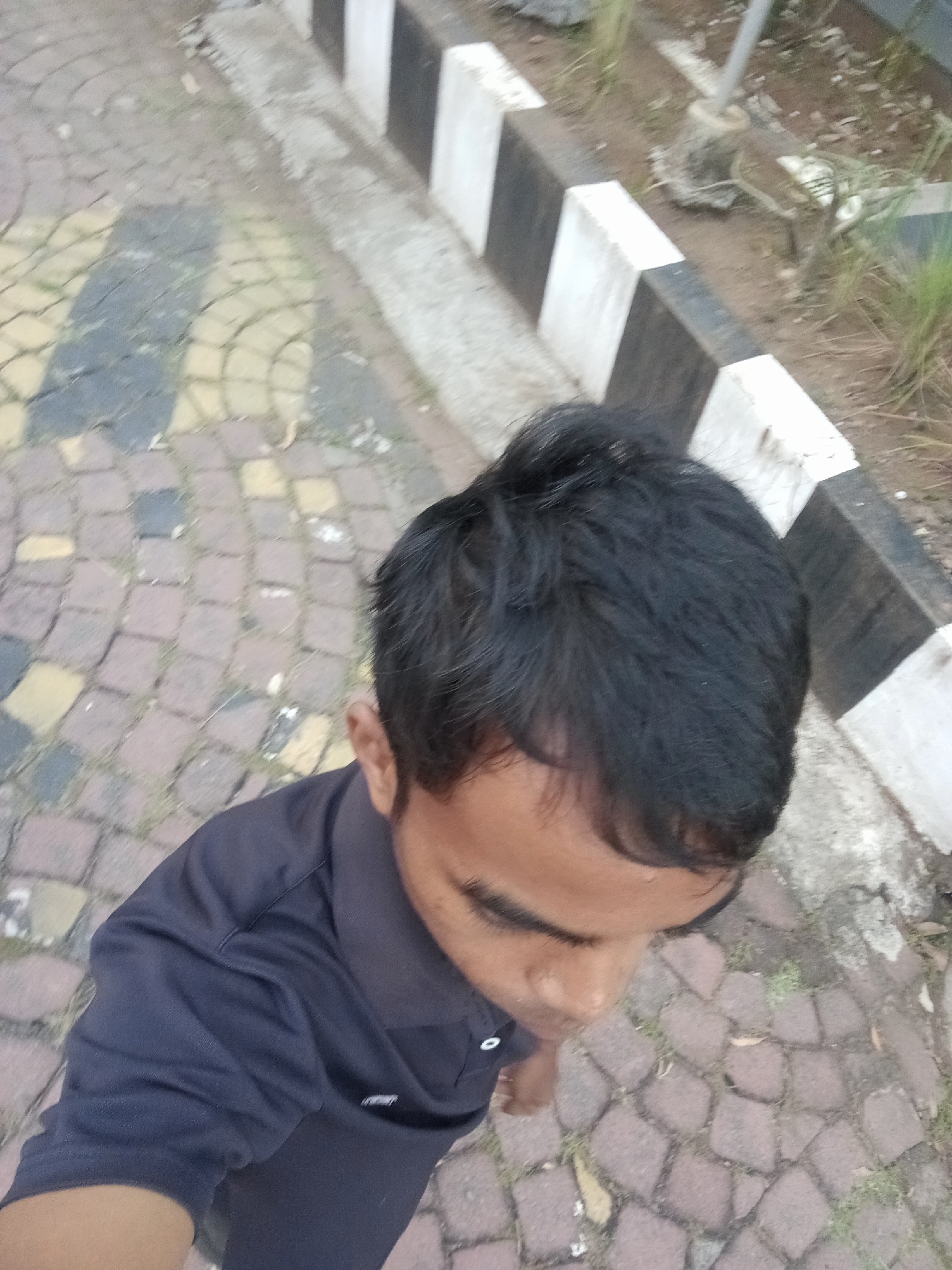 The image shows a close-up, high-angle shot of a person's head and upper body. The person has dark hair and is wearing a dark blue collared shirt. They are looking downwards and to the right. The background consists of a patterned stone walkway and a curb with alternating black and white blocks. The walkway appears to be outdoors, and there is some dry vegetation and dirt visible beyond the curb. The overall lighting suggests it is daytime. Based on the typical paving style and the curb design, this location is consistent with public walkways found in various areas of Surabaya, Indonesia, such as parks, pedestrian zones, or areas surrounding public facilities.