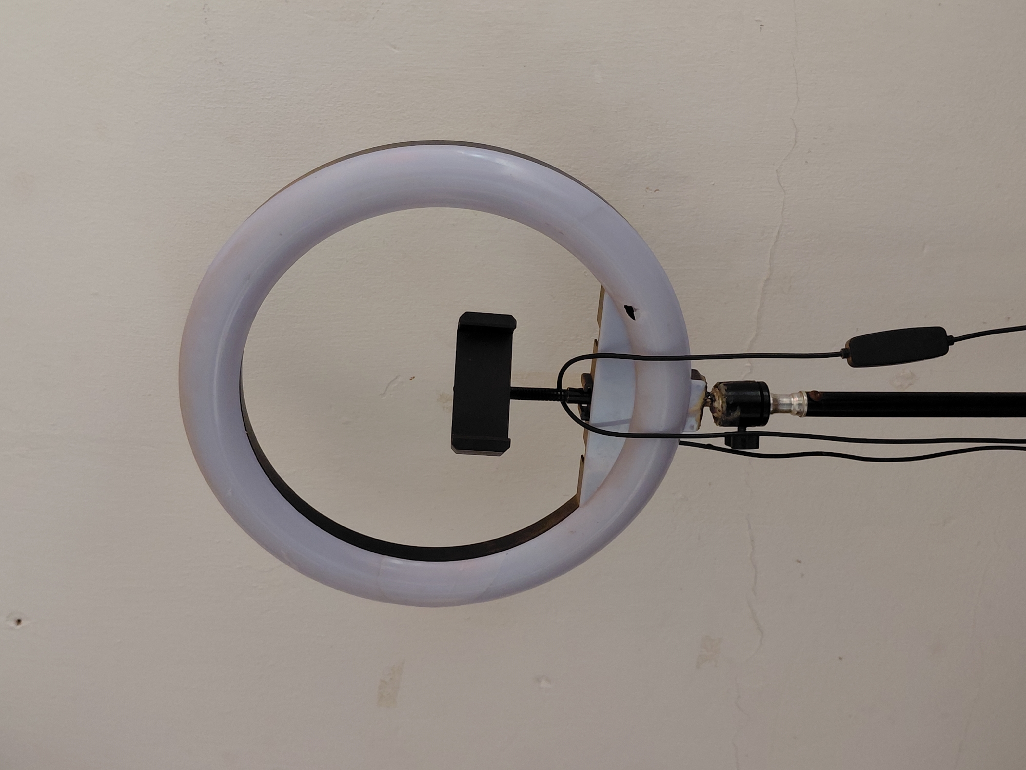 An image captured from a low angle shows a white ring light with a black phone holder positioned horizontally against a light-colored, textured wall. The ring light is circular, featuring a distinct black inner edge. A black phone holder is mounted centrally within the ring, suggesting its use for hands-free recording or streaming. On the right side, the ring light is connected to a black pole or stand. Black electrical cables run from the ring light along the pole, incorporating a black, rectangular control switch. The background wall, likely off-white or beige, exhibits signs of age or wear, including minor cracks and faint discoloration or dirt marks. No specific activity is depicted, nor are there discernible indications of time of day, weather, or emotions. No text is visible within the image. While the location context is Santa Ana, Venezuela, no visual elements in the image directly confirm this geographical setting.